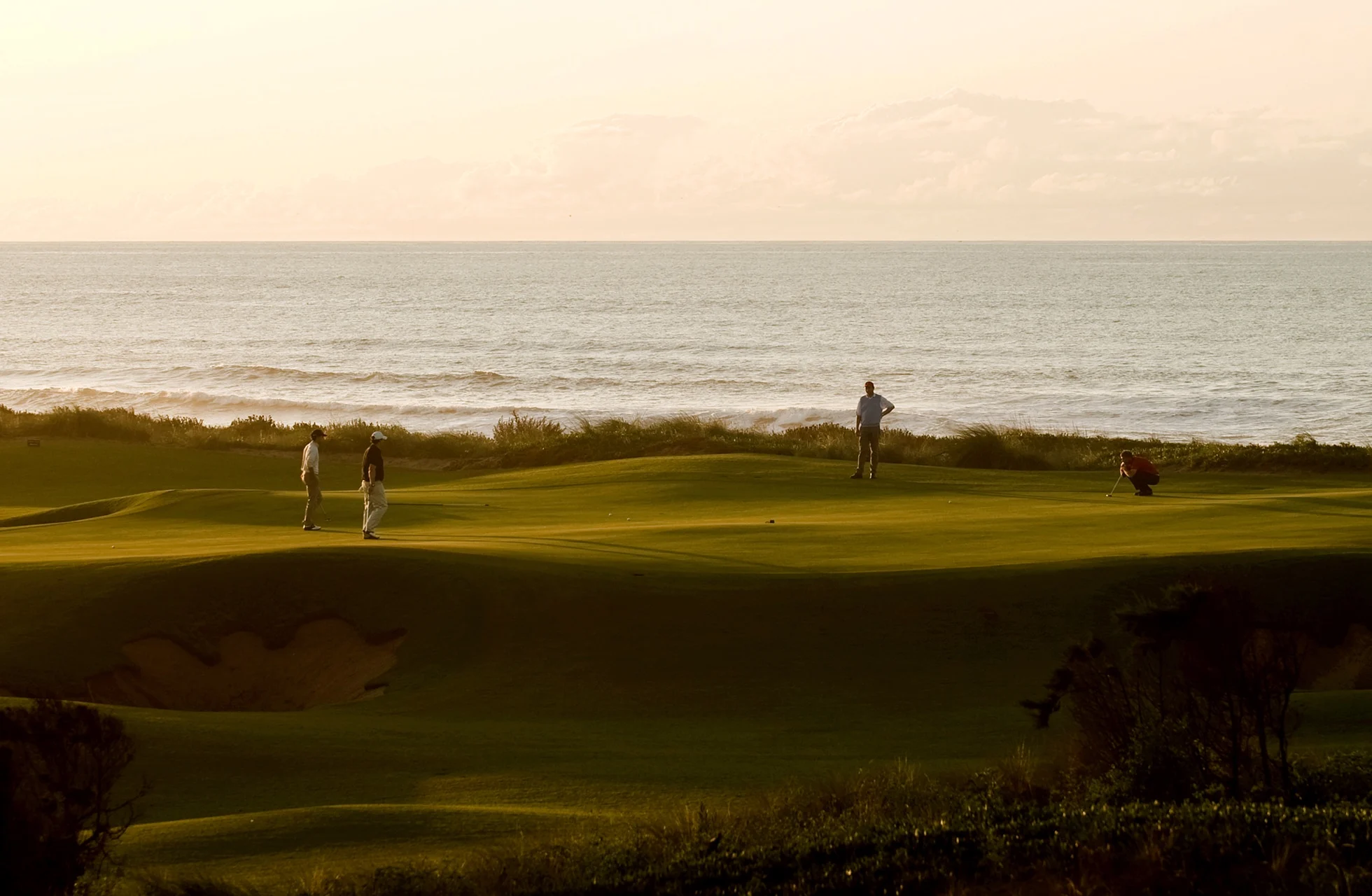 golfgreen i kystlandskap med flere golfspillere på greenen, havet i bakgrunnen og varmt lys fra sen ettermiddag eller kveld