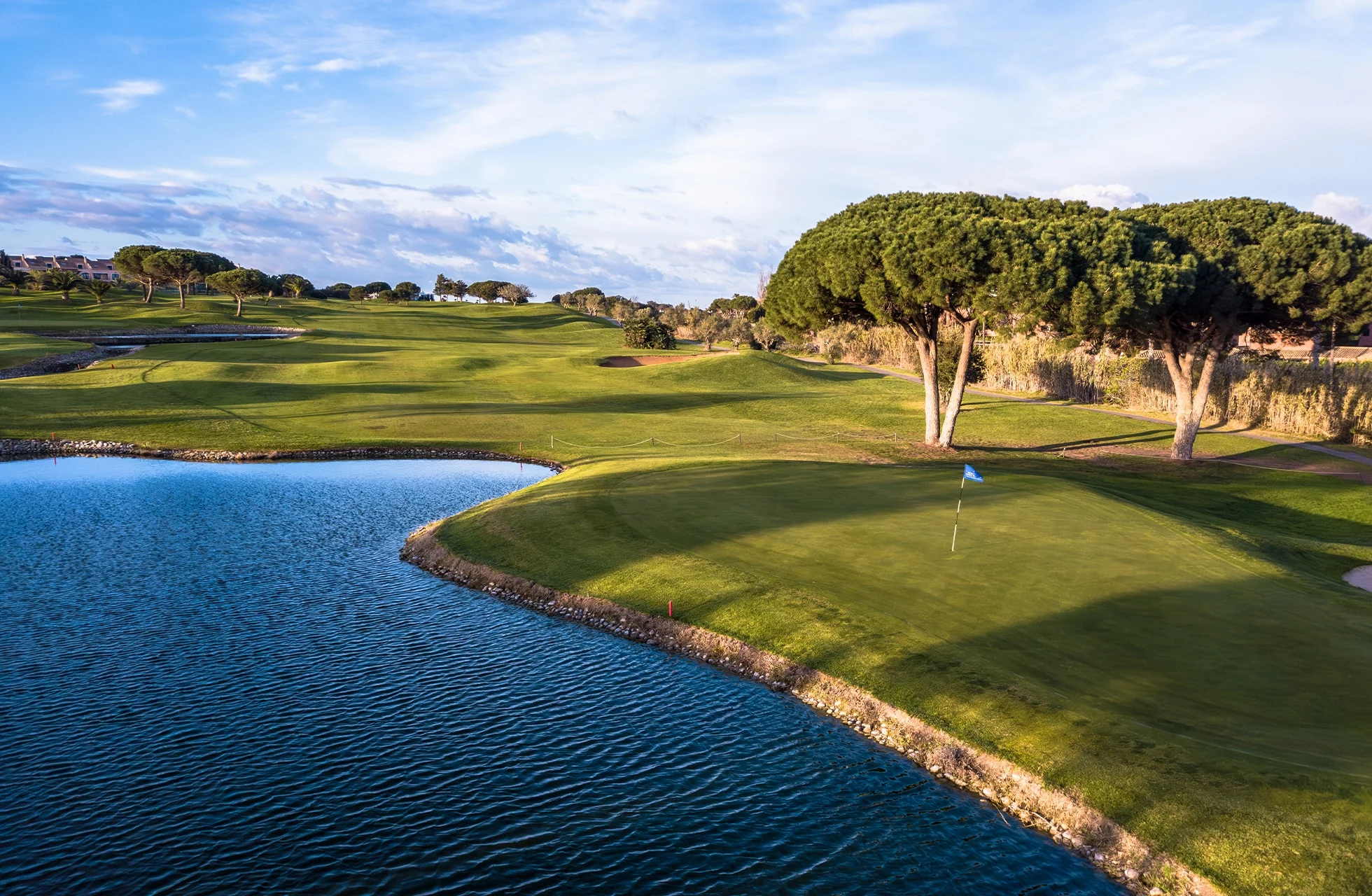 golfgreen med flagg ved et vannhinder, omgitt av bølgende fairwayer, bunkere og karakteristiske trær i kveldslys