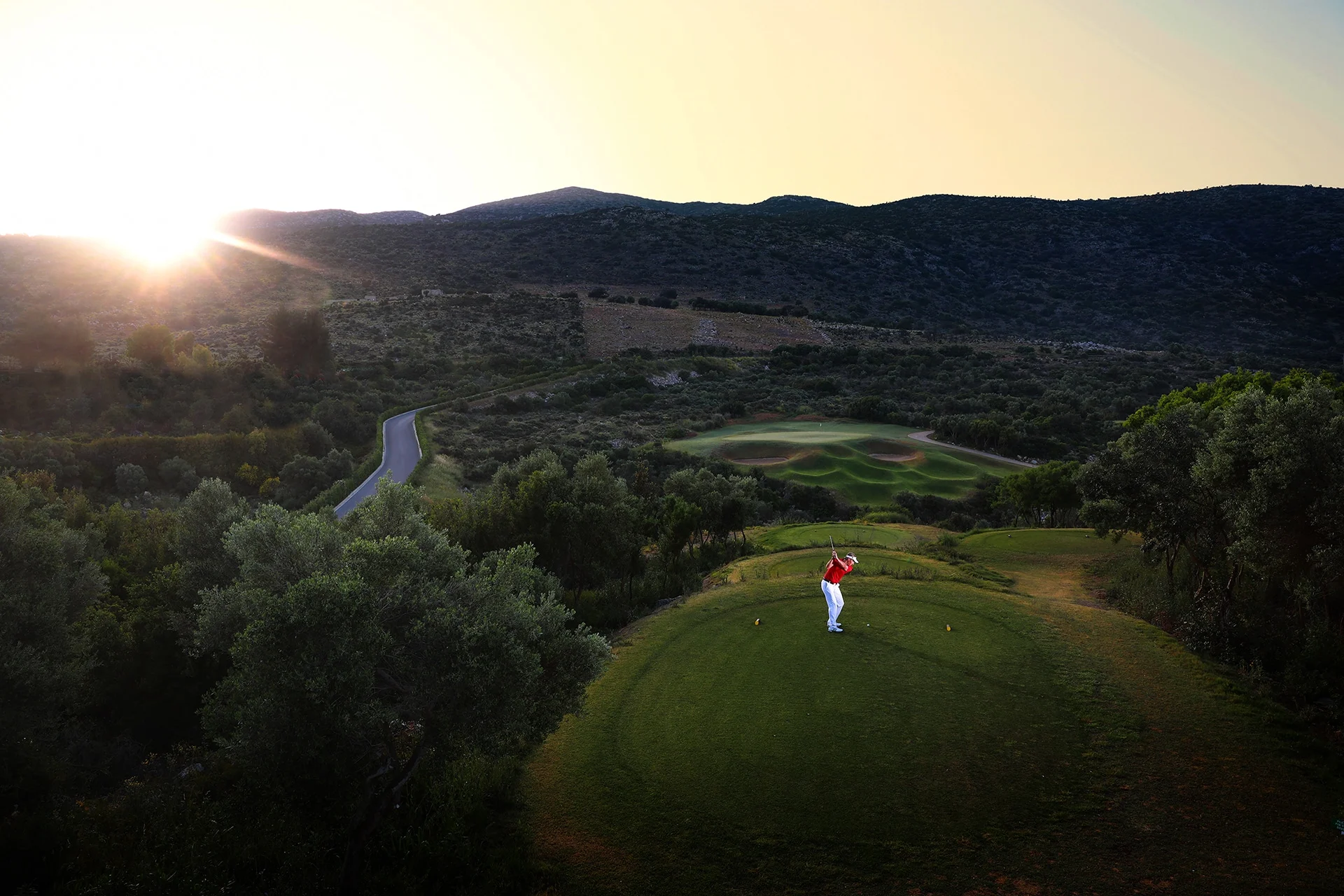 golfspiller slår ut fra tee i solnedgang, omgitt av trær, åser og svingende golfbane.