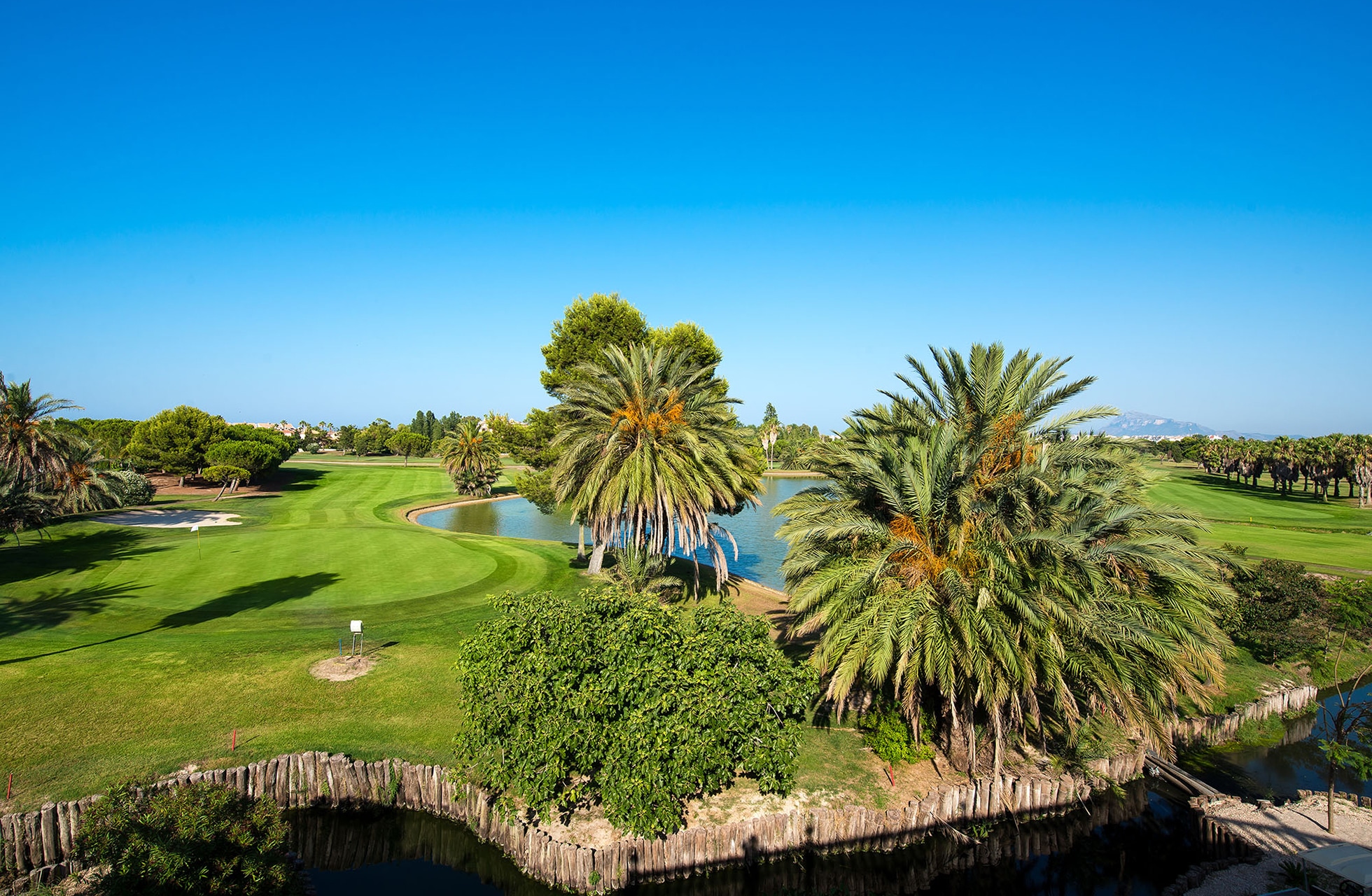 grønn golfbane med velklipte fairwayer og green, vannhinder i forgrunnen og høye palmer rundt banen under klar blå himmel i dagslys