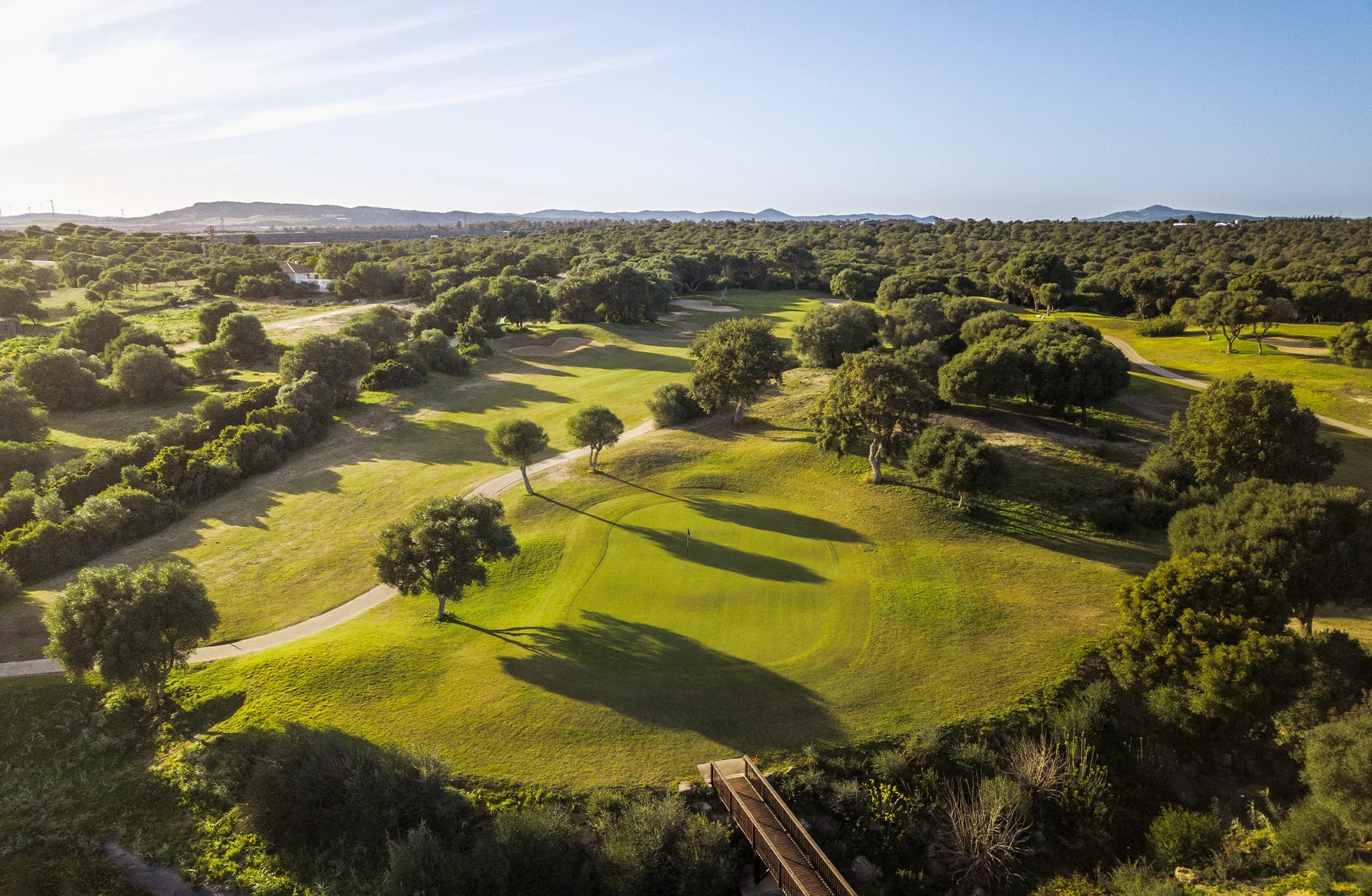 Panoramautsikt over golfbane med bølgende fairways, trær og mykt kveldslys sett ovenfra.
