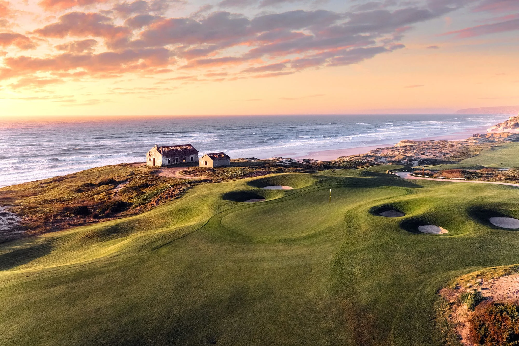 kystnær golfbane på praia d’el rey med greener og bunkere i bølgende landskap, atlanterhavet og kystlinjen i bakgrunnen i varmt kveldslys