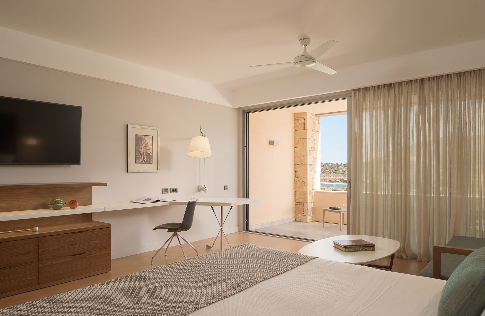lys juniorsuite med dobbeltseng, skrivebord og balkongdør som leder ut til terrasse i varme naturtoner.