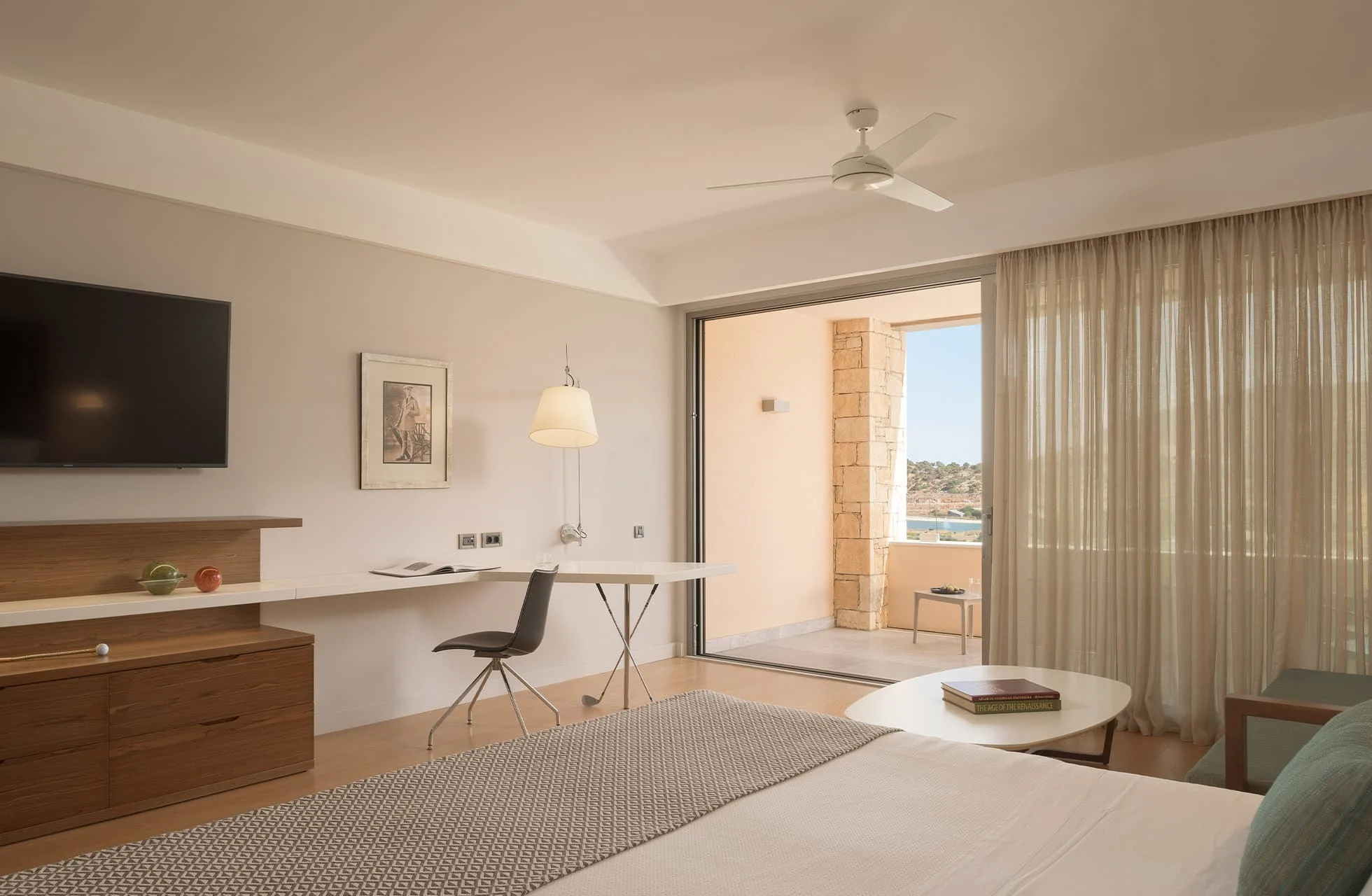 lys juniorsuite med dobbeltseng, skrivebord og balkongdør som leder ut til terrasse i varme naturtoner.