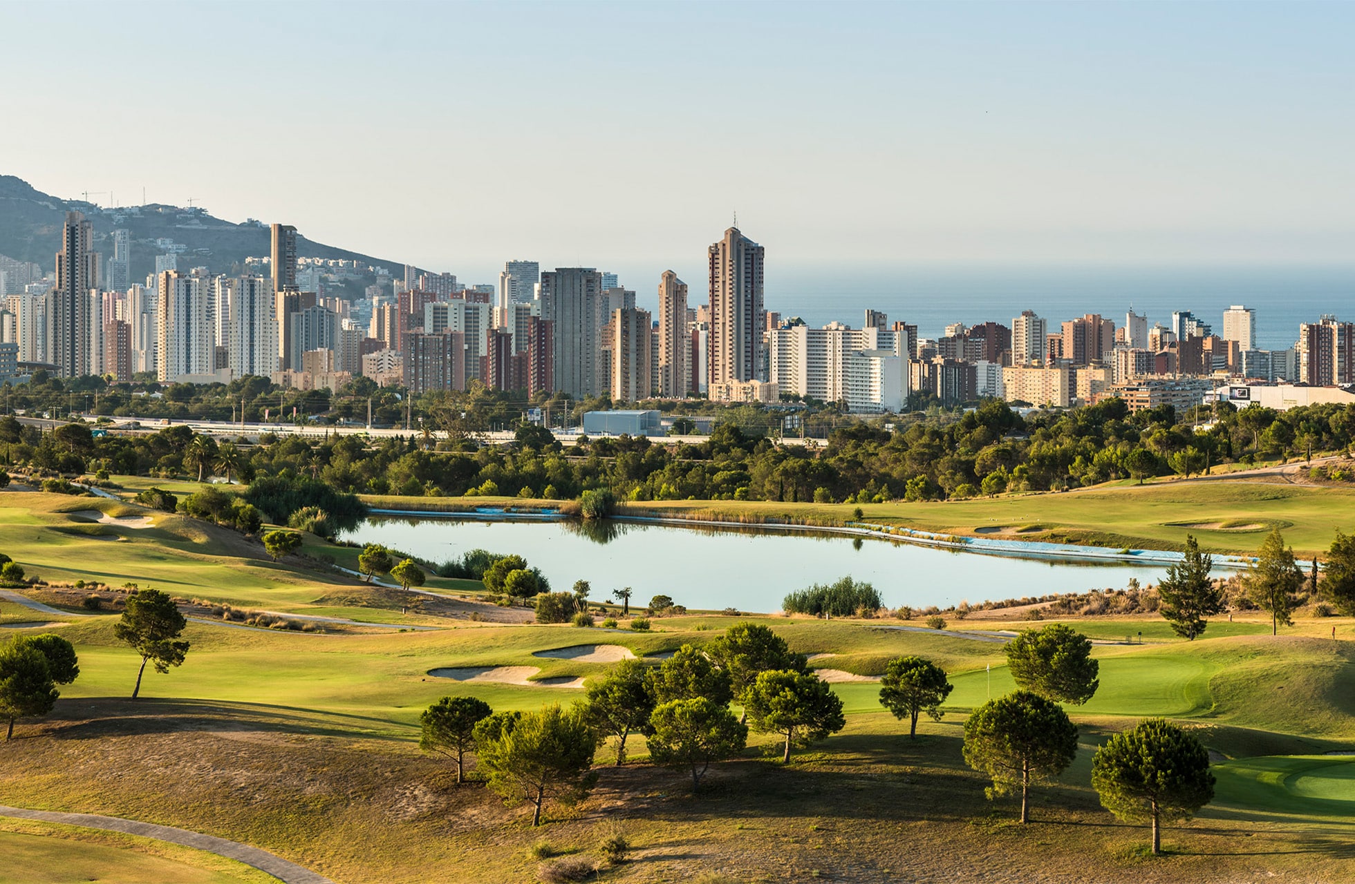 Oversikt over golfbanen med innsjøer, greener og skyline i bakgrunnen.
