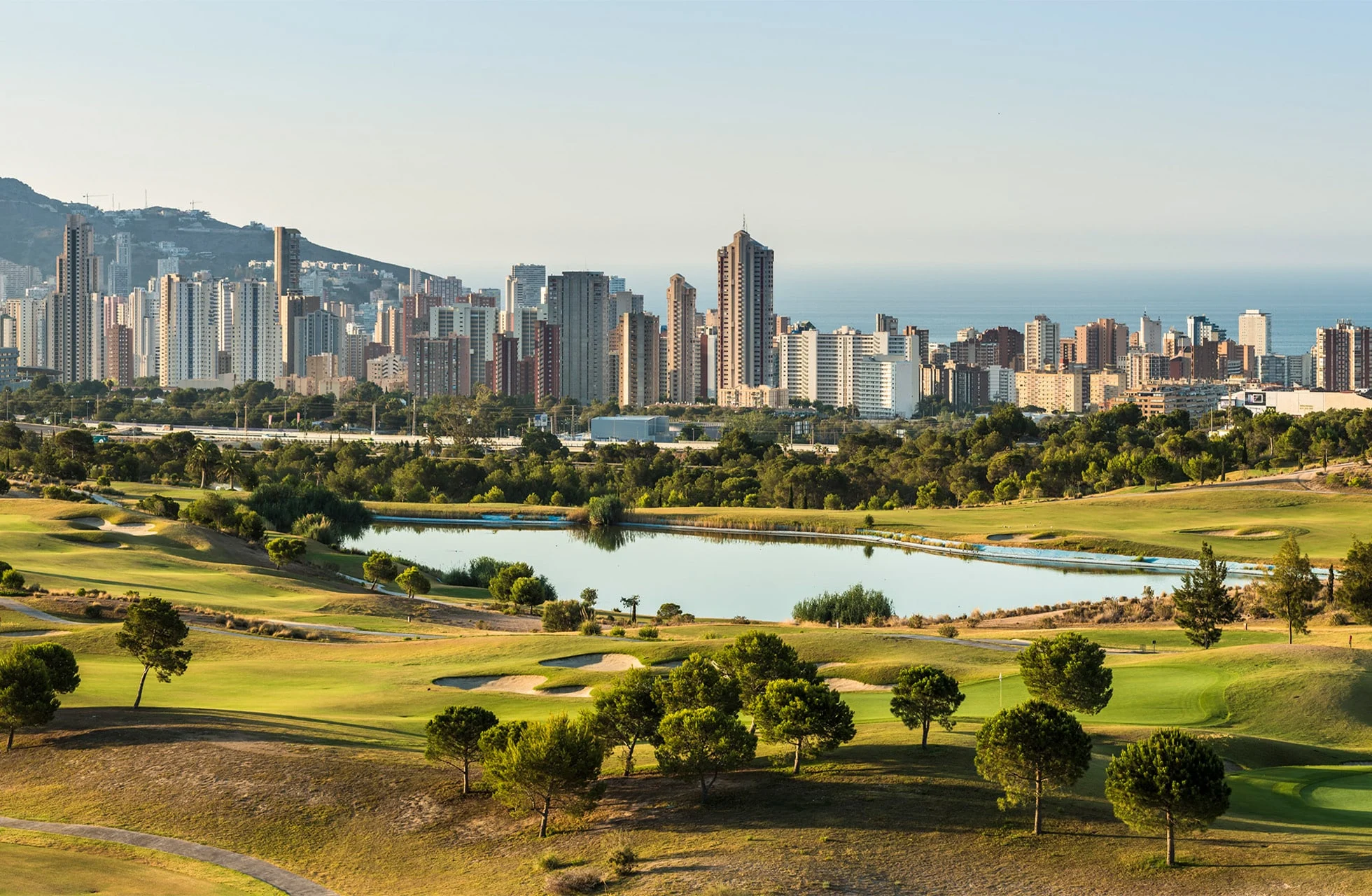 Oversikt over golfbanen med innsjøer, greener og skyline i bakgrunnen.
