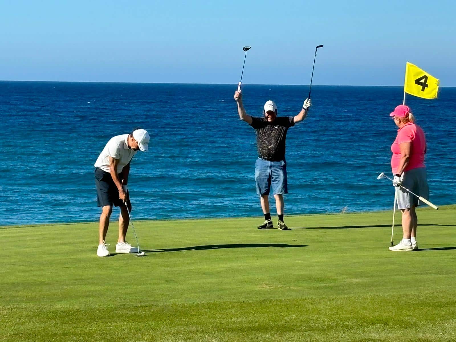 tre personer på en golfgreen ved havet, én putter mens to andre står ved siden av, med flagg merket med tallet 4