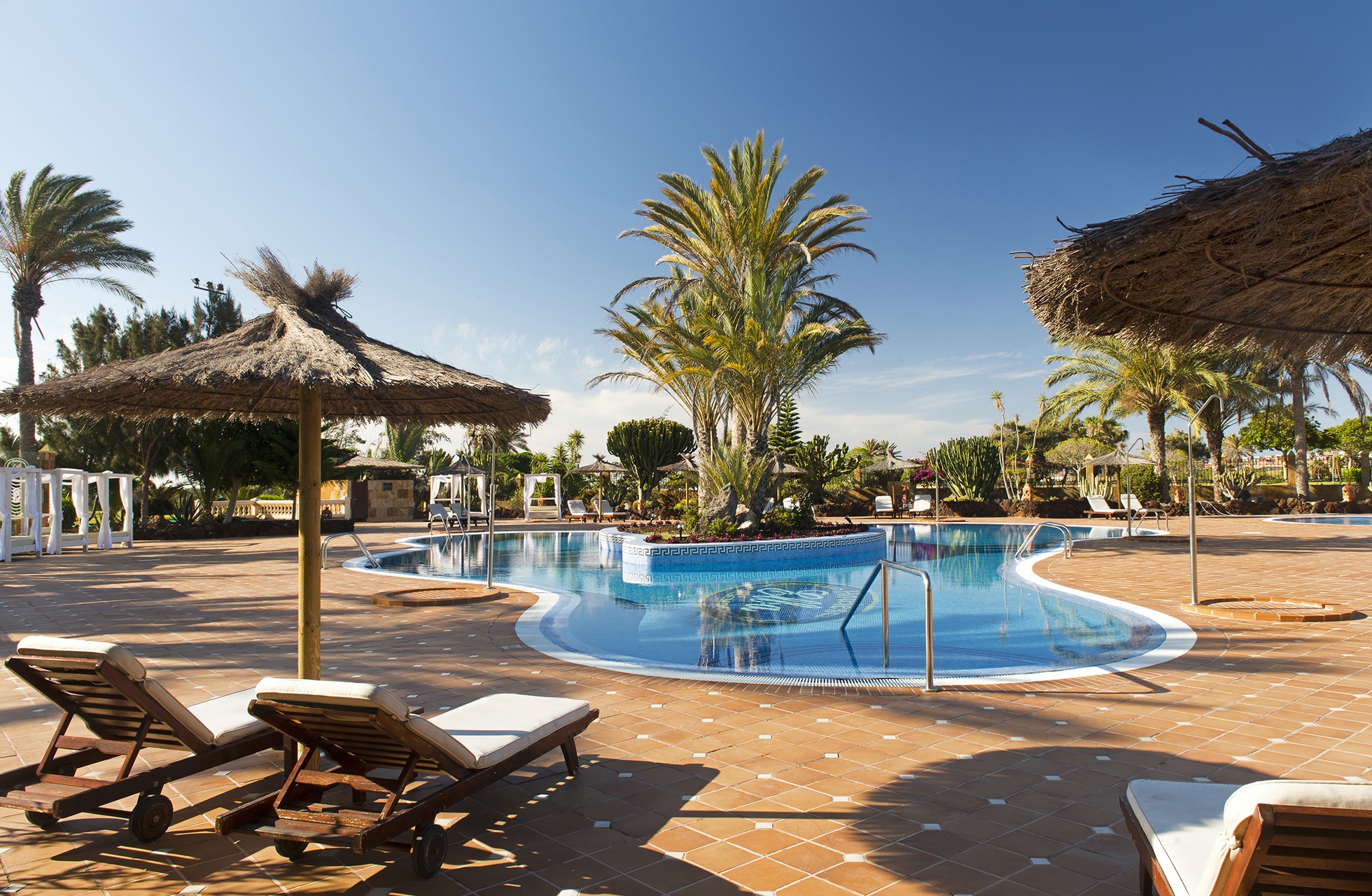 utendørs basseng på elba palace golf med rund pool, solsenger, stråparasoller, palmer og grøntområder i dagslys