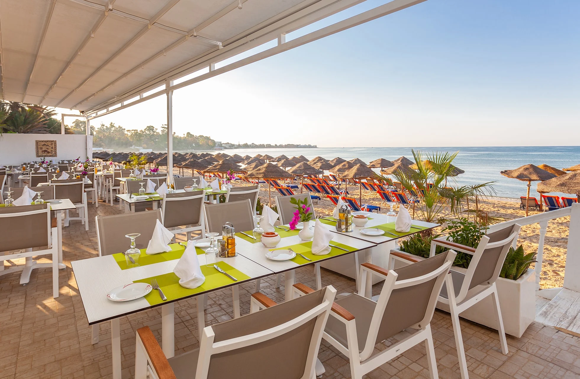 dekkede bord på åpen restaurantterrasse med stoler og fargerike duker, utsikt mot strand med solsenger og stråparasoller samt havet i bakgrunnen i klart dagslys
