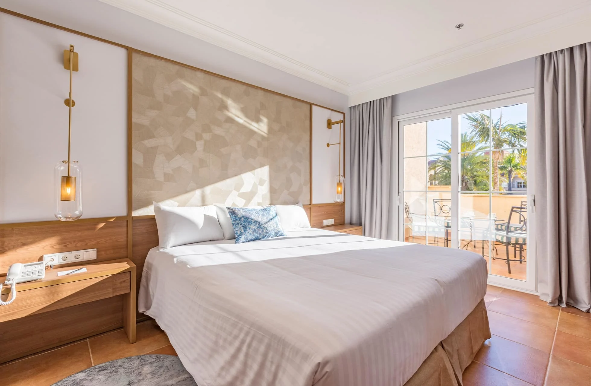 moderne hotellrom med dobbeltseng, nattbord og veggmonterte lamper, stort vindu og glassdør til balkong med utemøbler og palmer utenfor i dagslys