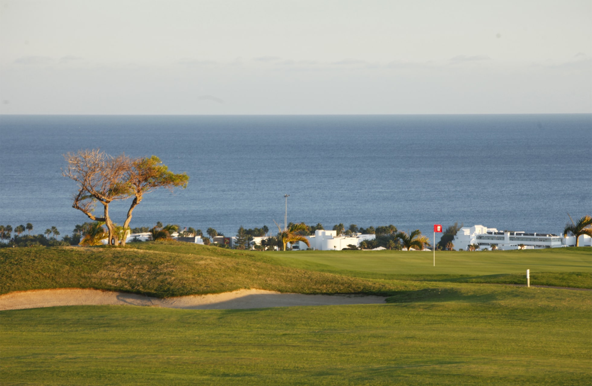 golfgreen med bunker i förgrunden och flagga på hålet. bakom greenen syns låga byggnader, träd och havet vid horisonten under en ljus himmel.