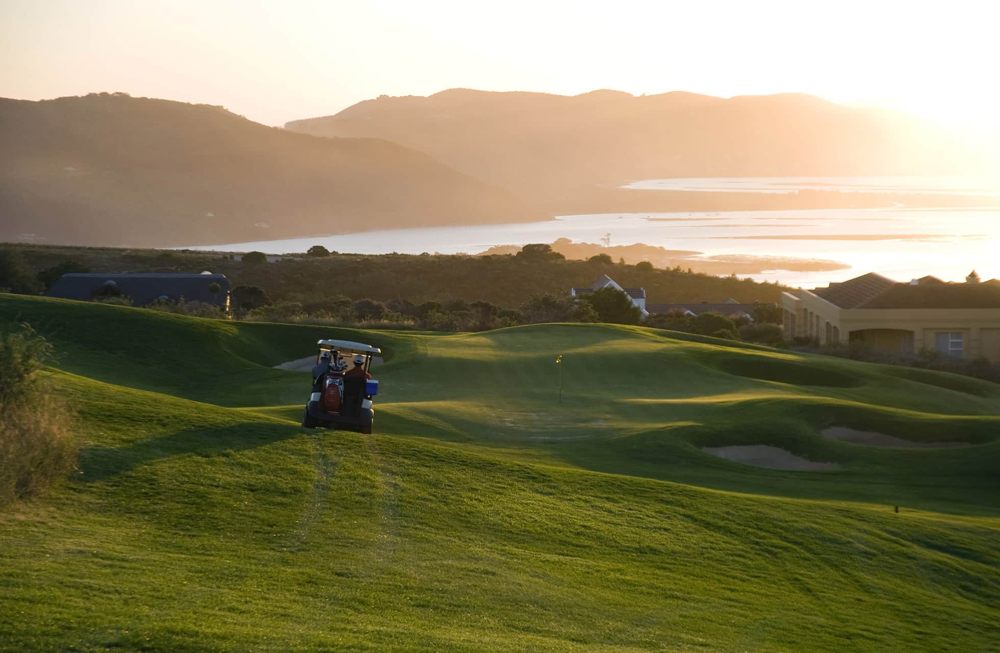 udsigt over en grøn golfbane i kuperet landskab med en golfvogn på fairway, omgivet af bygninger, bjerge og vand i varmt aftenslys