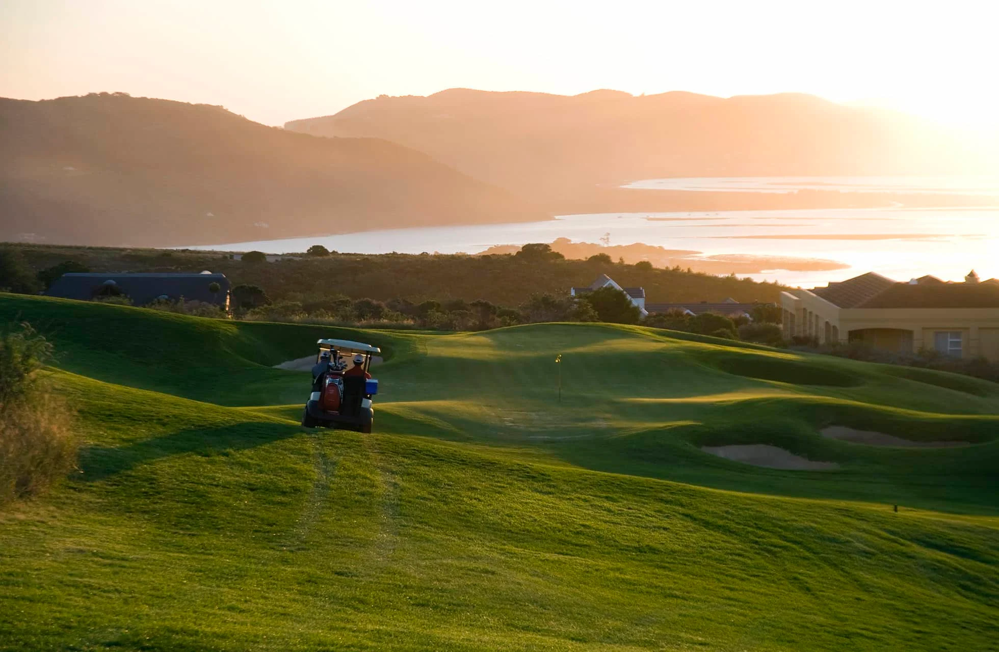 udsigt over en grøn golfbane i kuperet landskab med en golfvogn på fairway, omgivet af bygninger, bjerge og vand i varmt aftenslys