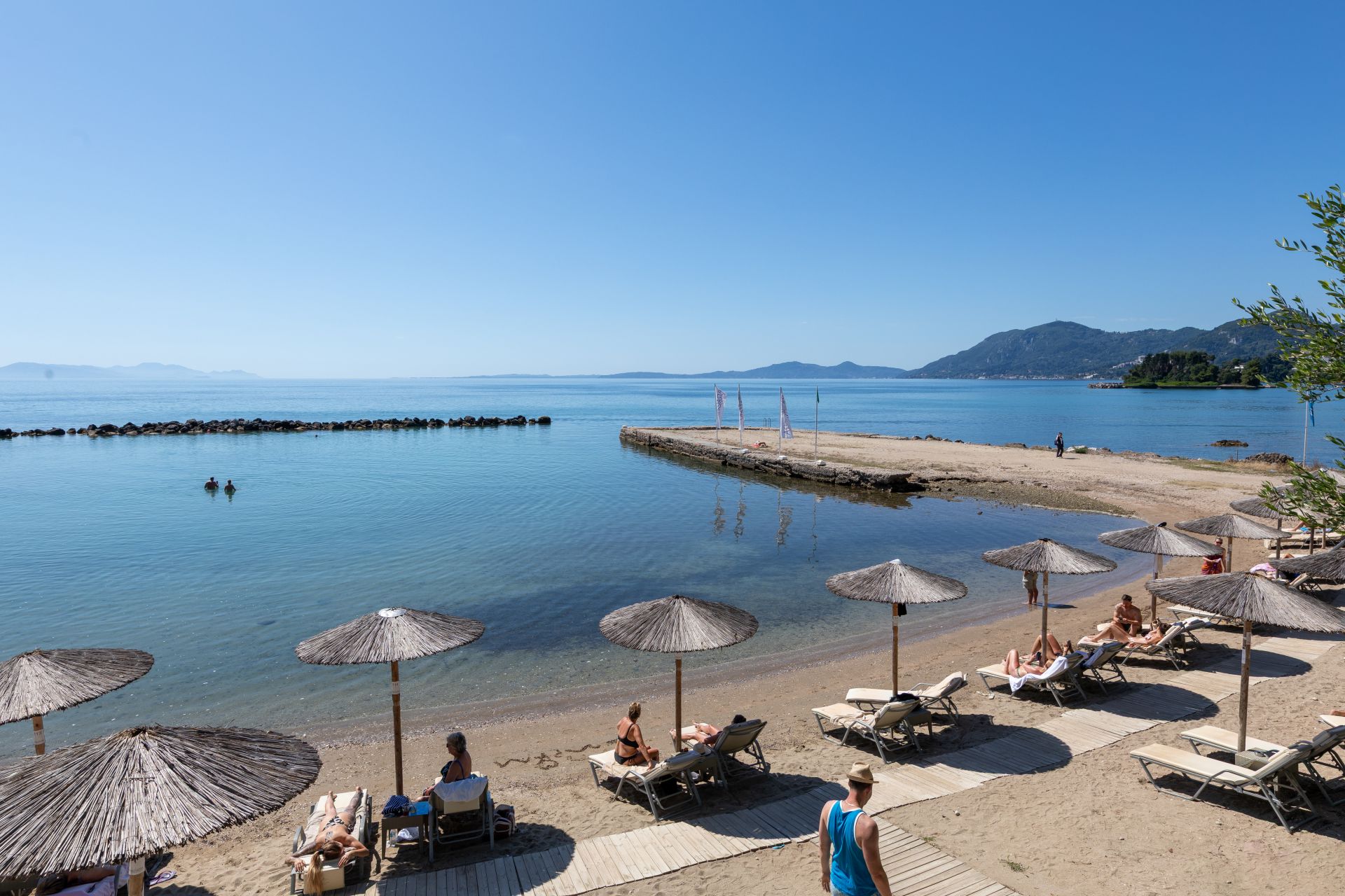 strand med parasoll och solstolar vid lugnt hav, pir och berg i bakgrunden