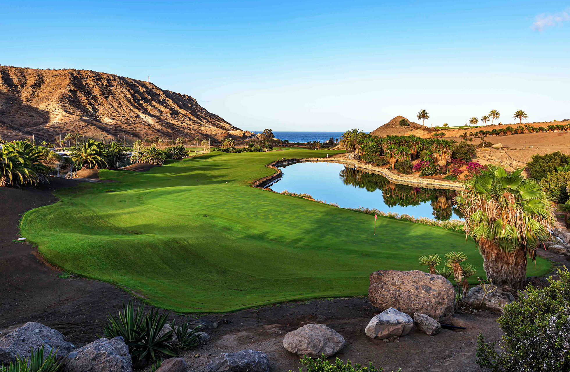 golfgreen på anfi tauro golf med vannhinder ved greenen, fairway i kupert landskap samt fjell og hav i bakgrunnen i klart dagslys