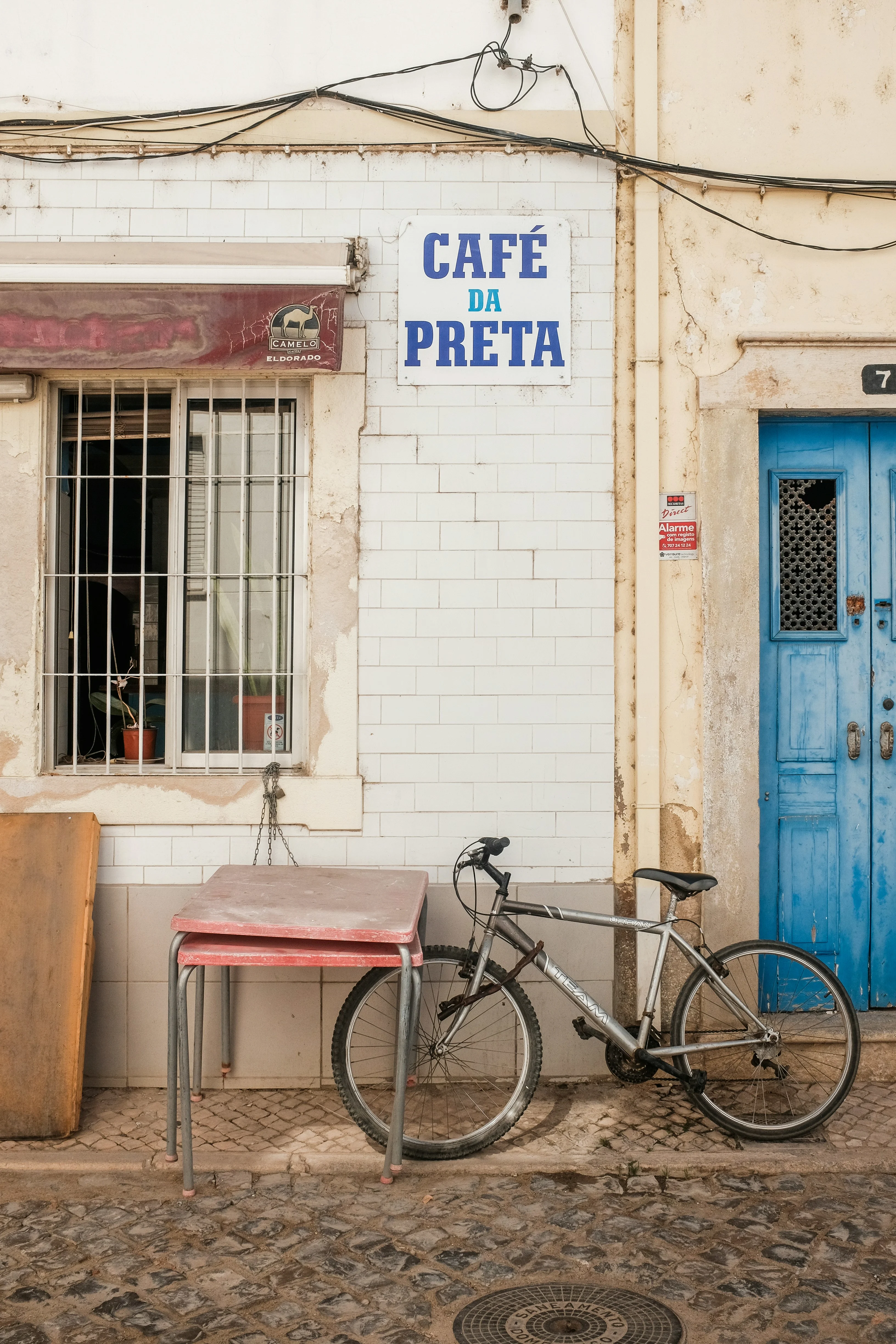 en sykkel står lent mot en lys mur utenfor et kafé med blå dør og et enkelt bord ved siden av