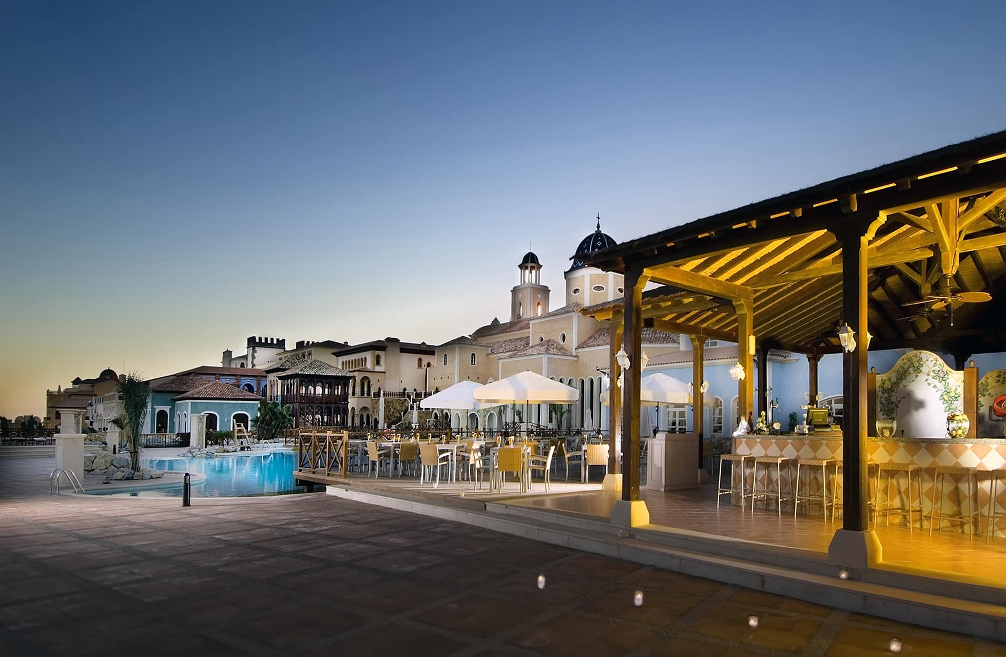 udendørs restaurant med dækkede borde under pergola ved en oplyst pool, flisebelagt terrasse og bygninger med tårne i baggrunden i aftenskumring
