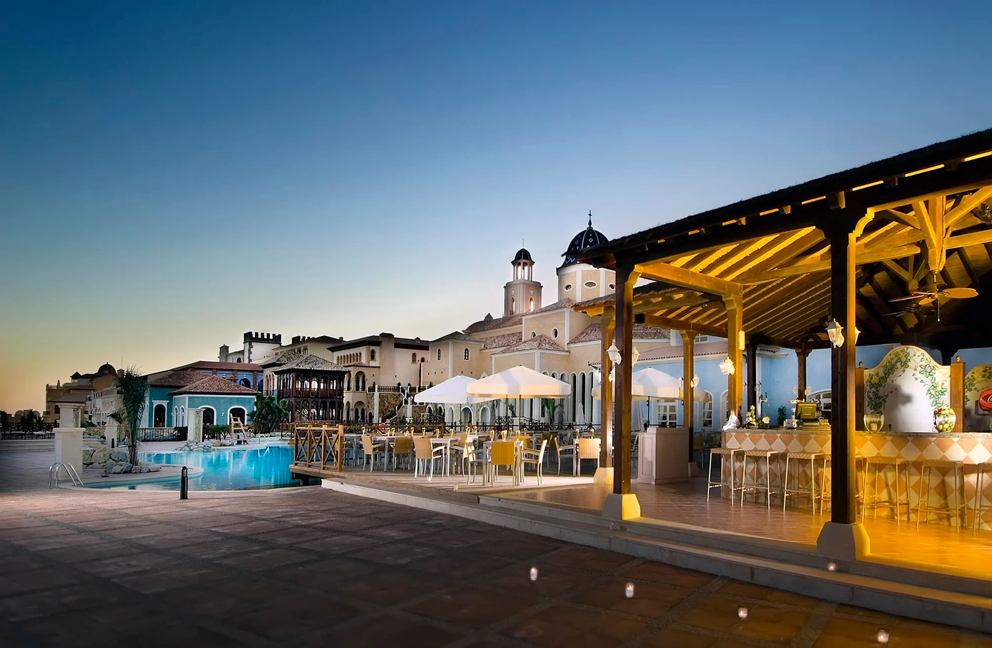udendørs restaurant med dækkede borde under pergola ved en oplyst pool, flisebelagt terrasse og bygninger med tårne i baggrunden i aftenskumring