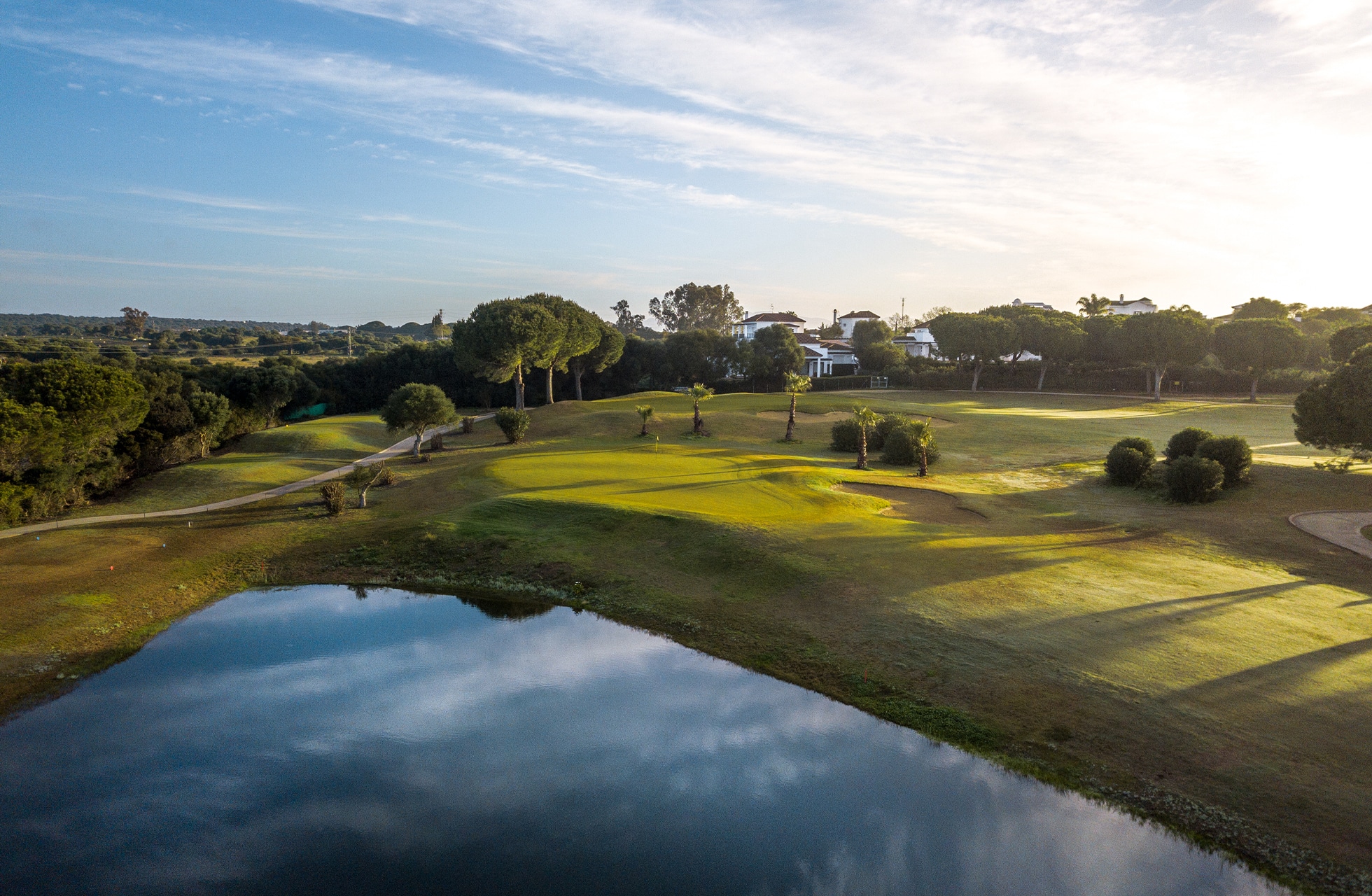 golfbane med vannhinder i forgrunnen, green og fairway i åpent landskap med trær og bygninger i varmt lys