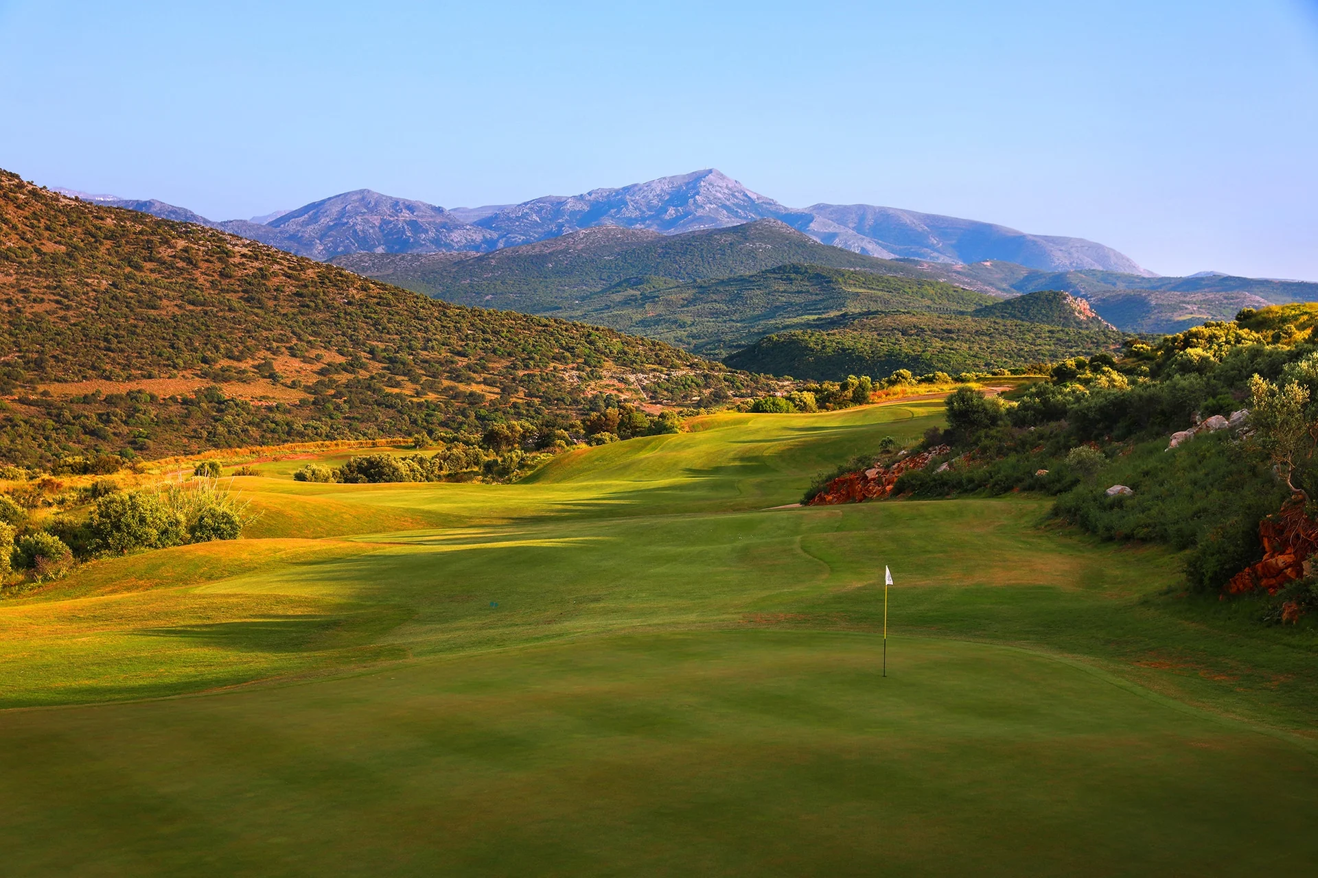 fairway og green som slynger seg gjennom åpent landskap med fjell i bakgrunnen og synlig flagg på greenen.