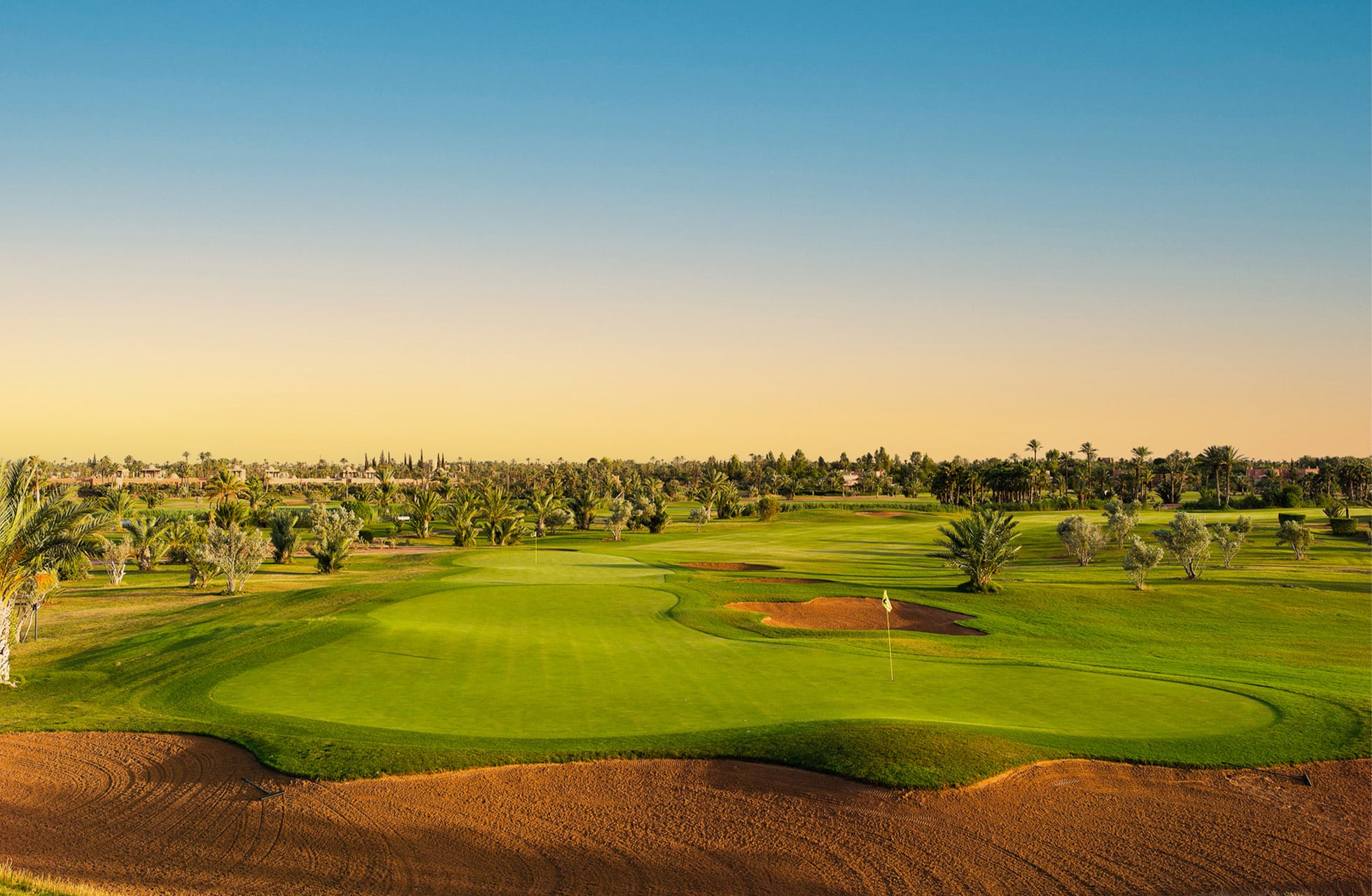 golfgreen med bunker i forgrunnen, omgitt av palmer og åpent landskap under klar himmel i varmt dagslys