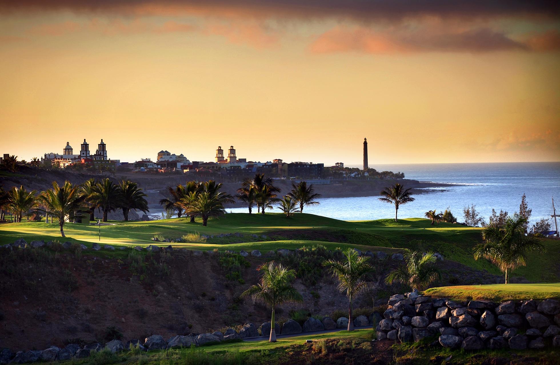 golfbane på meloneras golf i varmt kveldslys med green og palmer i forgrunnen, kystbyens silhuett og havet i bakgrunnen