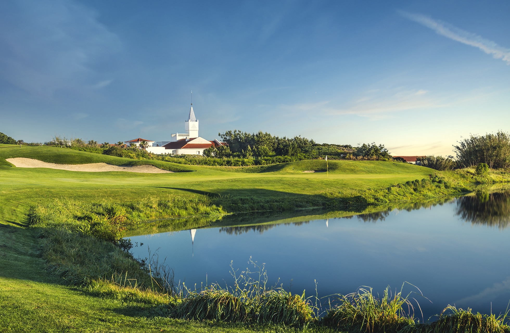 golfgreen med flagg i forgrunnen, speilblank dam foran greenen, bunkere og bølgende fairwayer, med bygninger og lav vegetasjon i bakgrunnen i klart dagslys