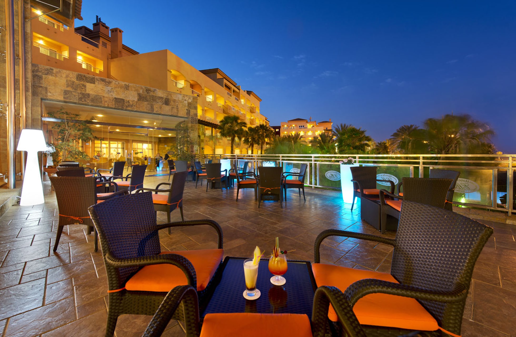 utendørs lounge på hotellterrasse med sittegrupper, serverte drinker, glassrekkverk og opplyst hotellmiljø i skumring