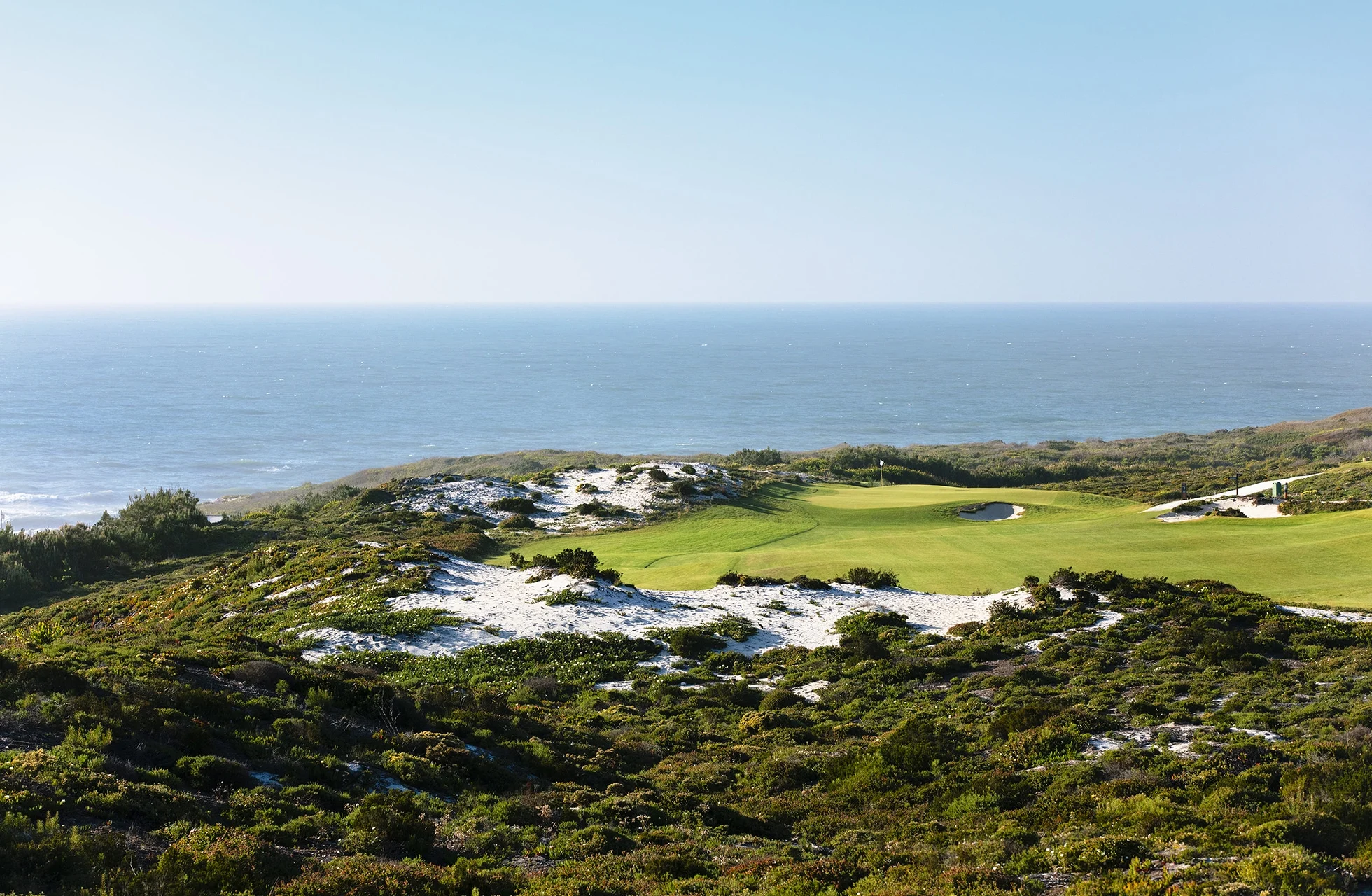 golfgreen omgitt av naturlig vegetasjon og sandpartier, med åpent hav synlig i bakgrunnen i klart dagslys