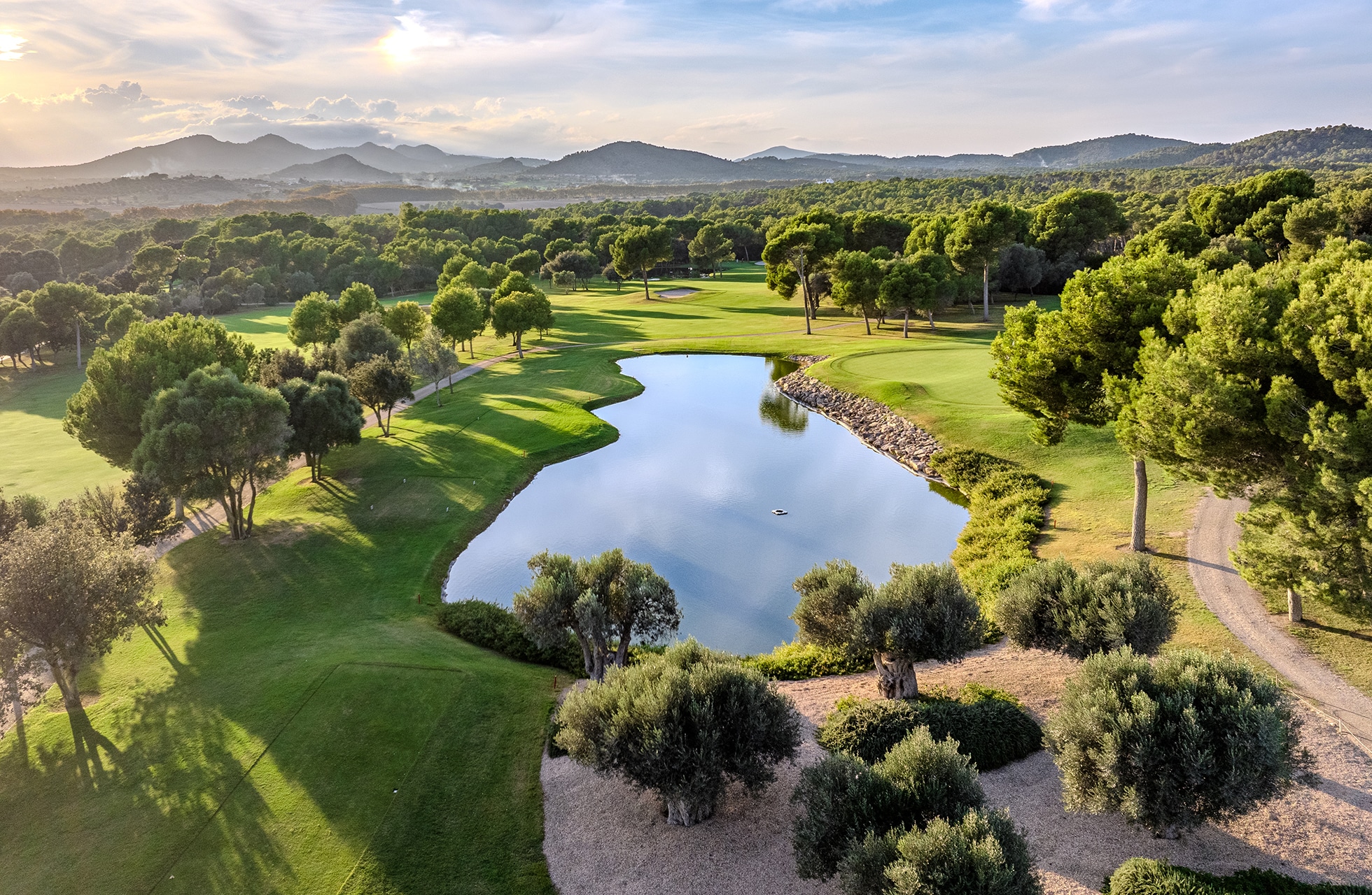 luftfoto av son servera golf med vannhinder, greener og fairwayer omgitt av furutrær og kupert landskap i mykt kveldslys.