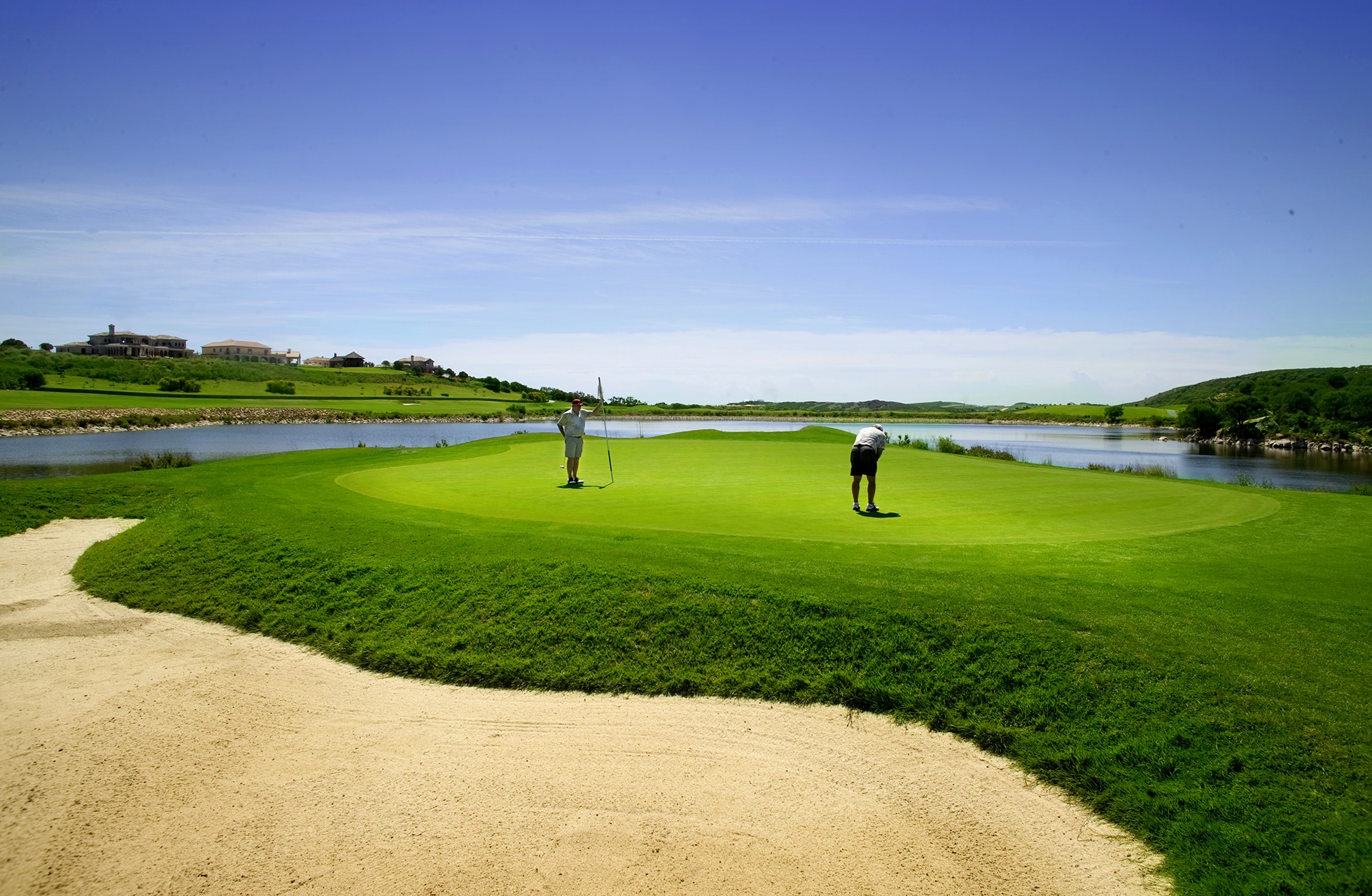 golfgreen med bunker i forgrunnen og vann rundt, to golfspillere som putter i åpent landskap i klart dagslys