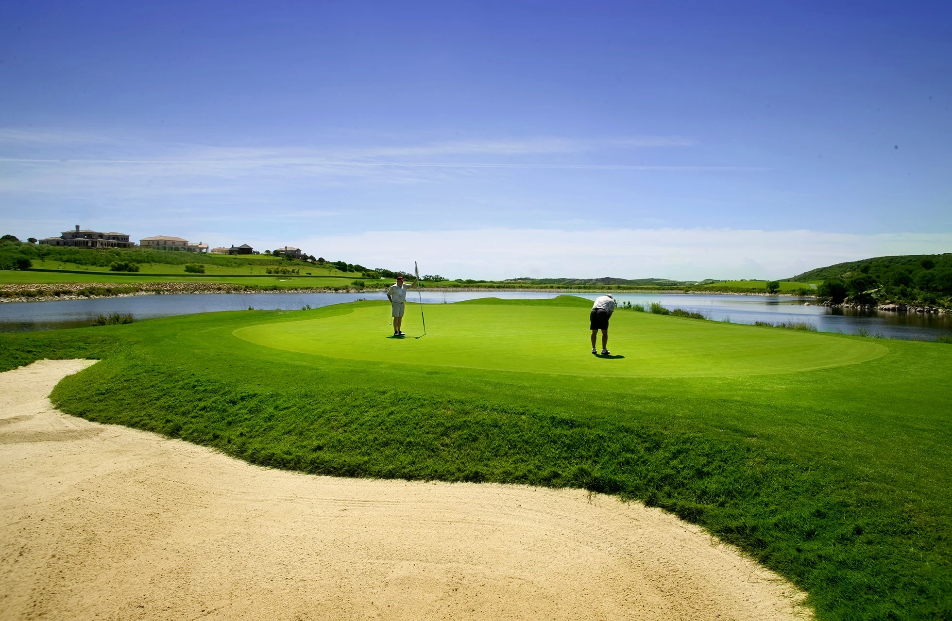 golfgreen med bunker i forgrunnen og vann rundt, to golfspillere som putter i åpent landskap i klart dagslys
