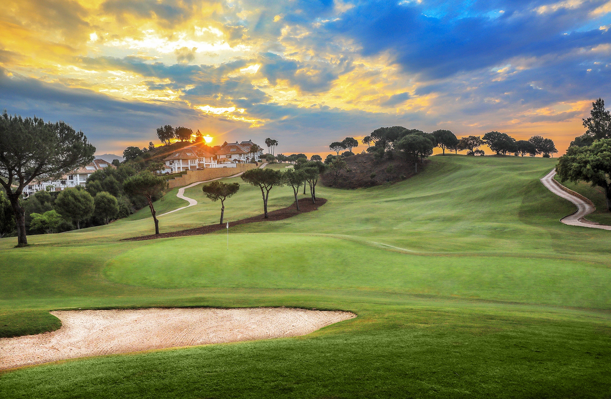 golfbane med bunker i forgrunnen, bølgende fairway og green i kupert landskap med trær og dramatisk himmel i kveldslys