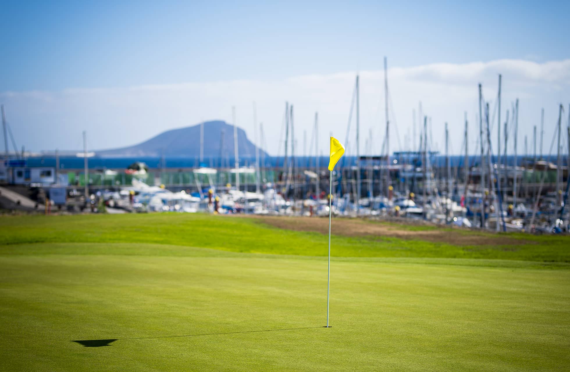 golfviheriö, etualalla lipputanko. viheriön takana näkyy venesatama purjeveneineen, mastoja ja rakennuksia sekä kauempana meri ja vuoret kirkkaan taivaan alla.