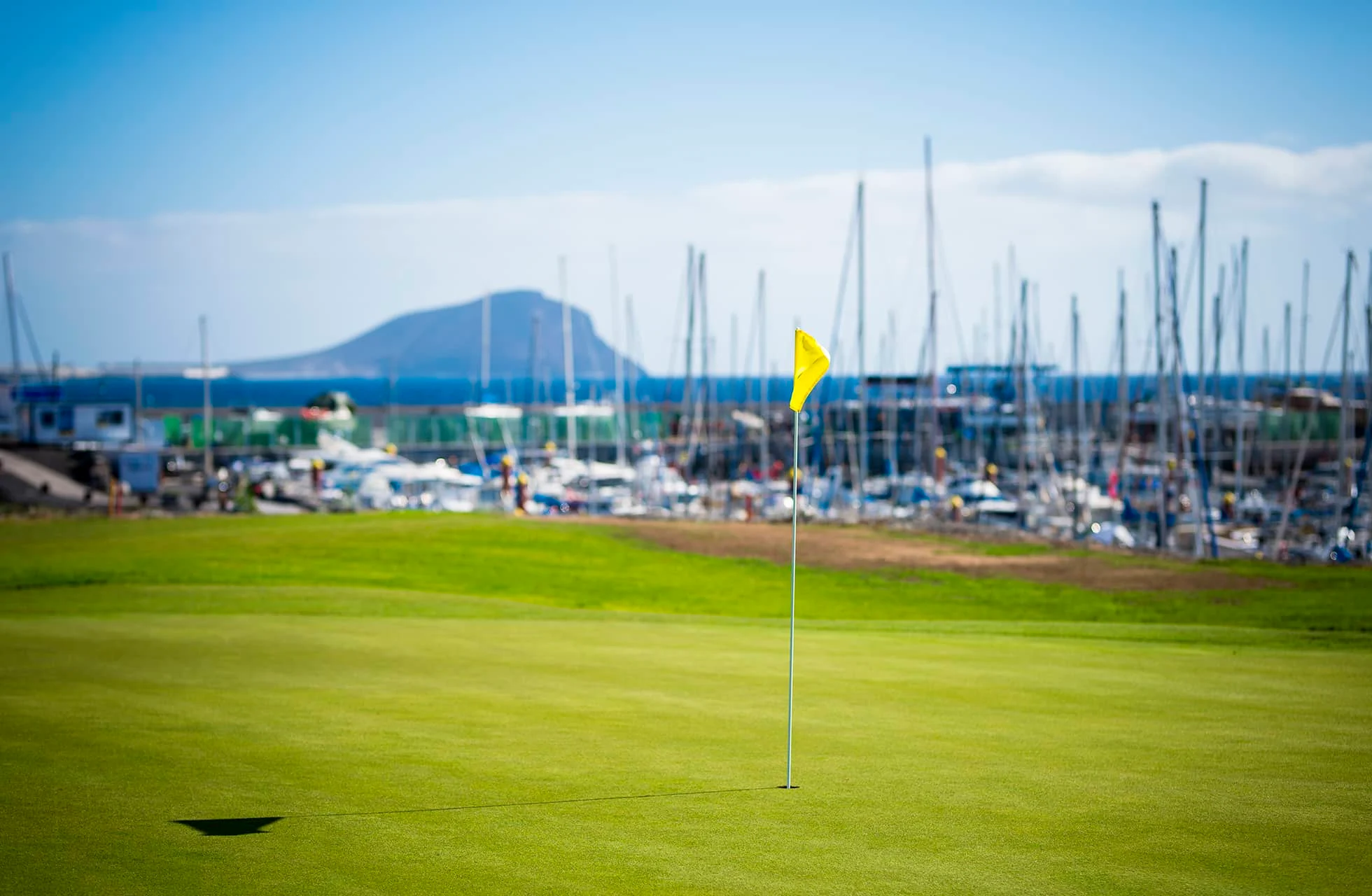 golfviheriö, etualalla lipputanko. viheriön takana näkyy venesatama purjeveneineen, mastoja ja rakennuksia sekä kauempana meri ja vuoret kirkkaan taivaan alla.