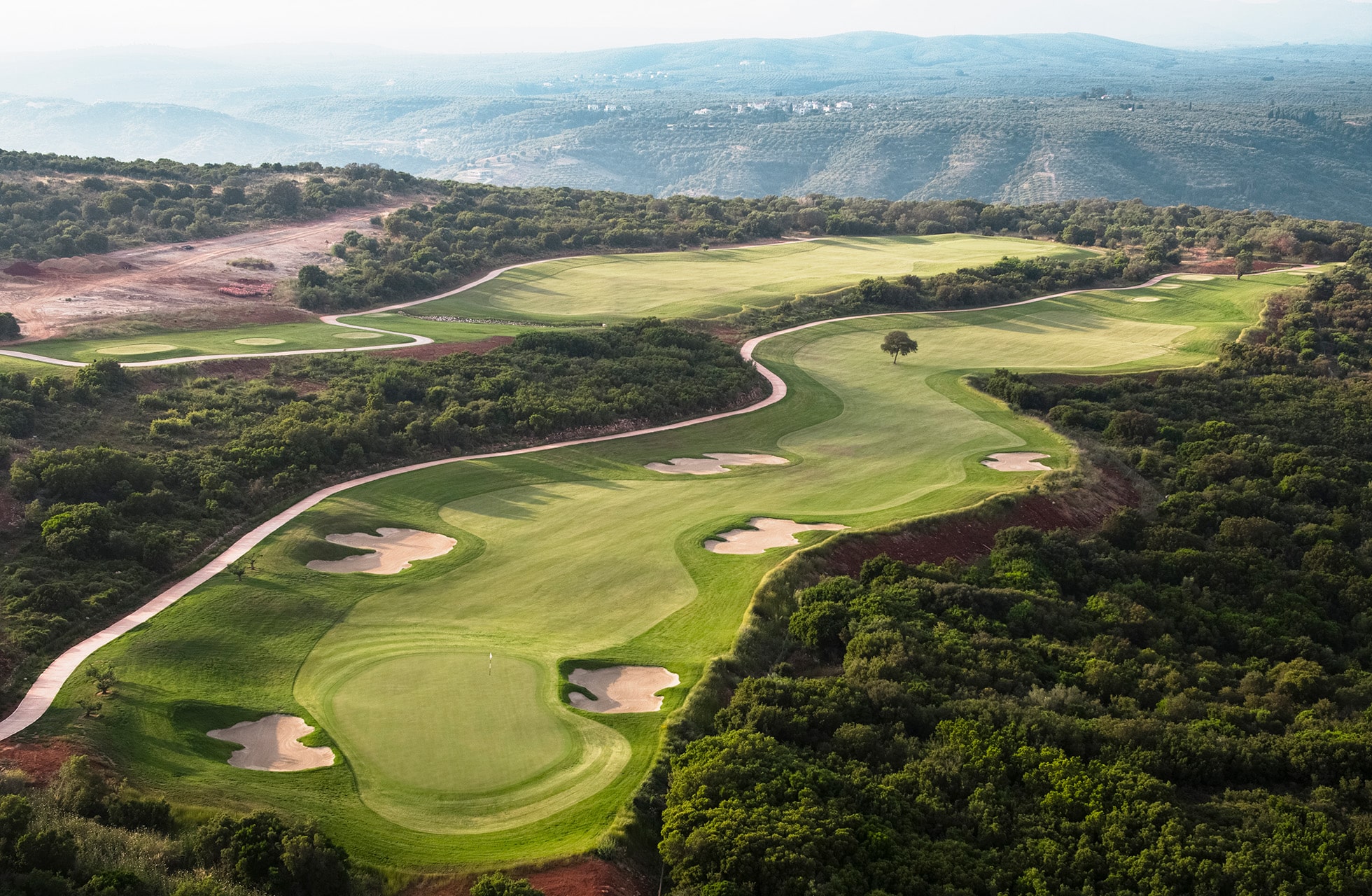utsikt over en grønn golfbane med fairwayer og bunkere som slynger seg gjennom kupert terreng med vegetasjon og fjell i bakgrunnen