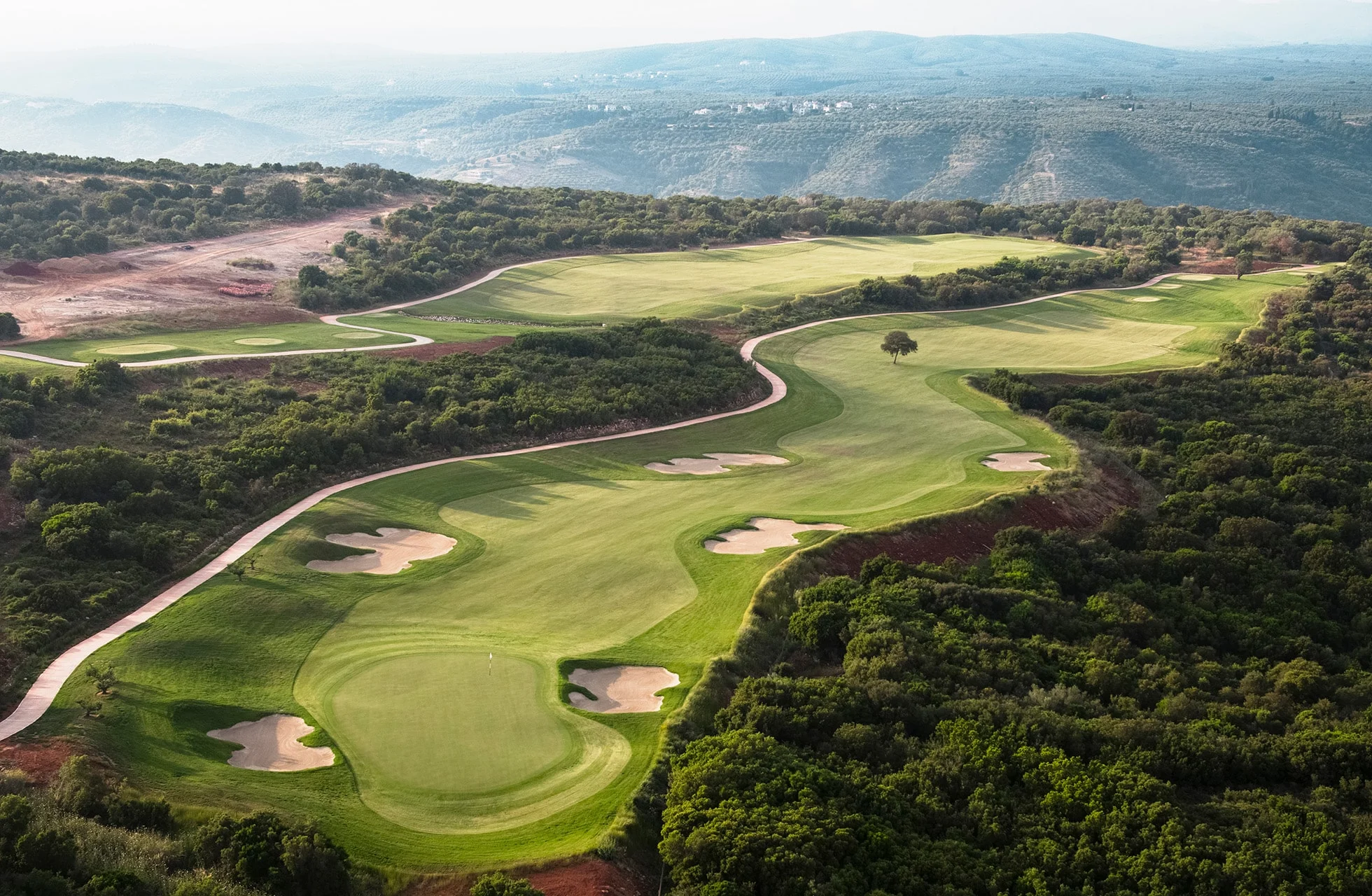 utsikt over en grønn golfbane med fairwayer og bunkere som slynger seg gjennom kupert terreng med vegetasjon og fjell i bakgrunnen