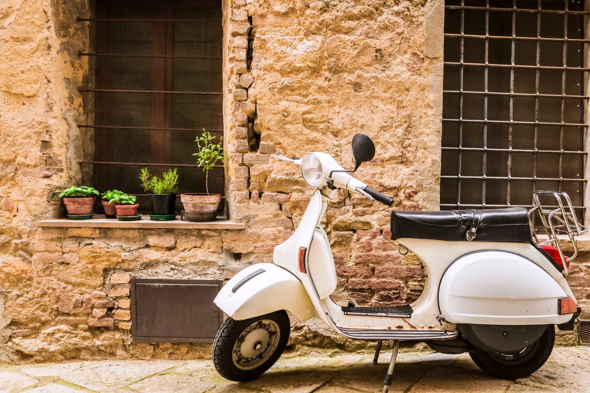 en hvid scooter parkeret foran en slidt stenmur med blomsterkrukker i vindueskarmen