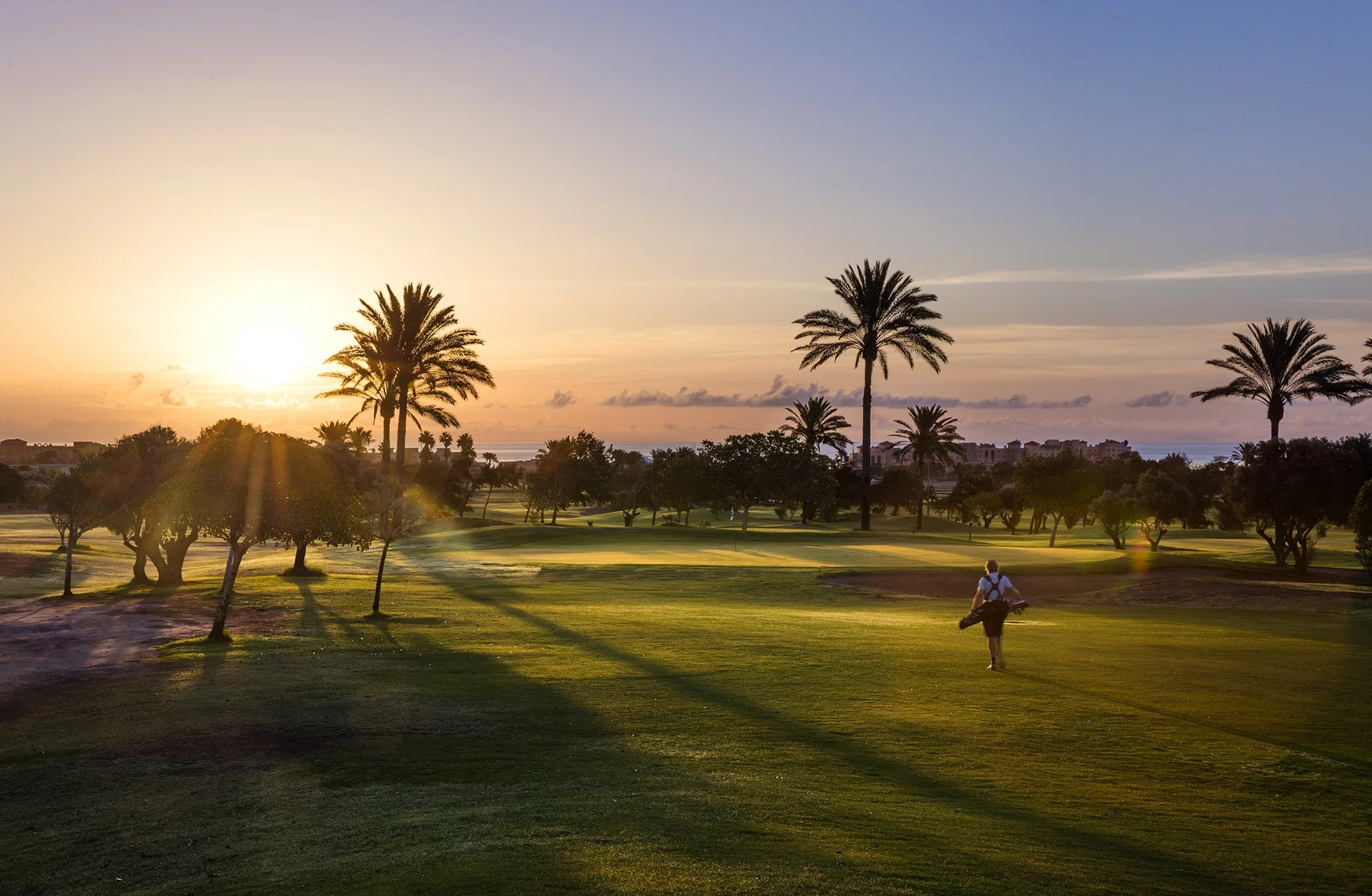 golfkenttä auringonlaskussa, jossa pitkät varjot ulottuvat väylälle. palmut ja puut kehystävät maisemaa, ja golfpelaaja kävelee nurmen yli lämpimässä iltavalossa.