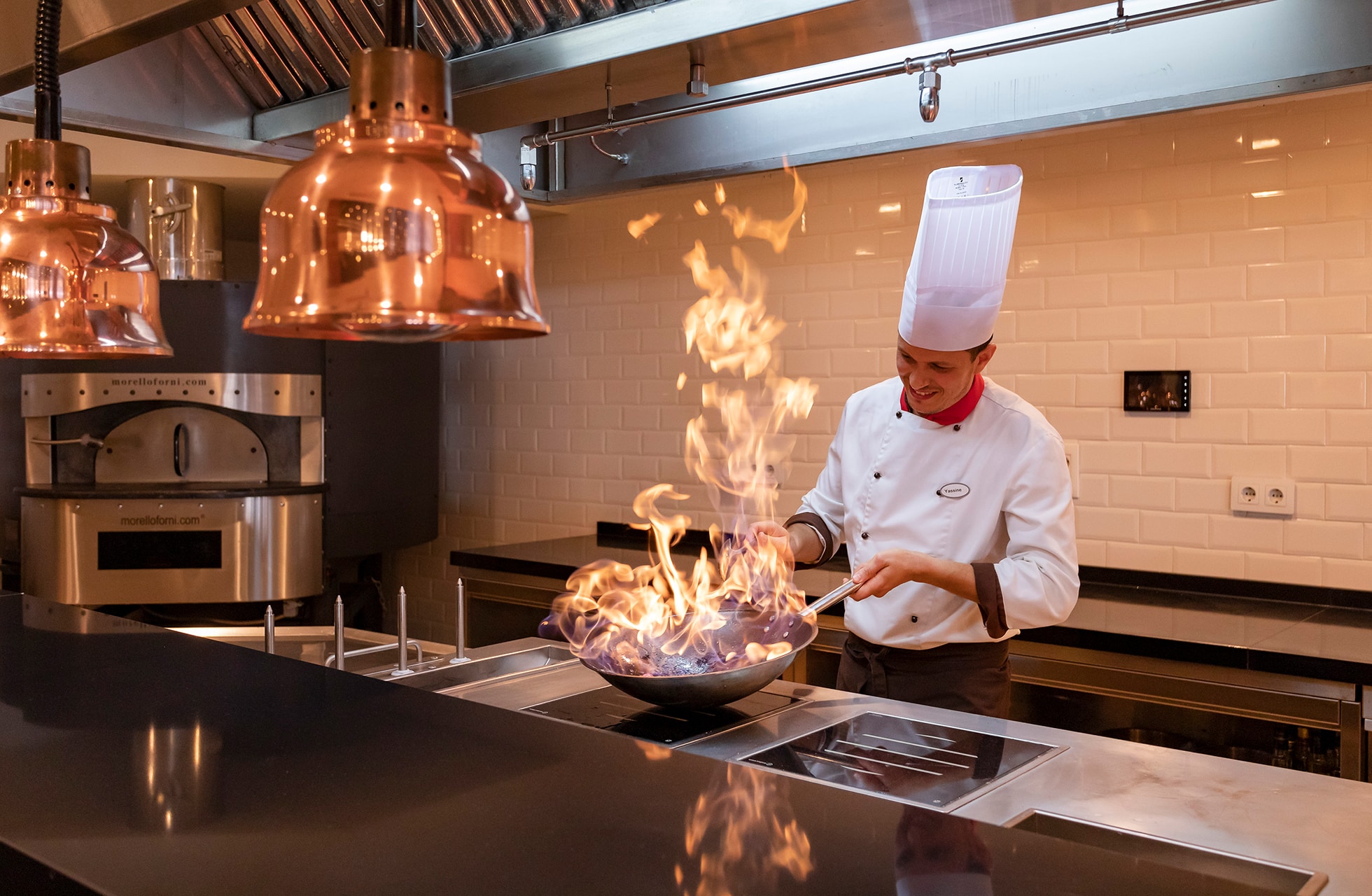 kokk i åpent restaurantkjøkken som lager mat i stekepanne med flammer, omgitt av profesjonelt kjøkkenutstyr og varm belysning