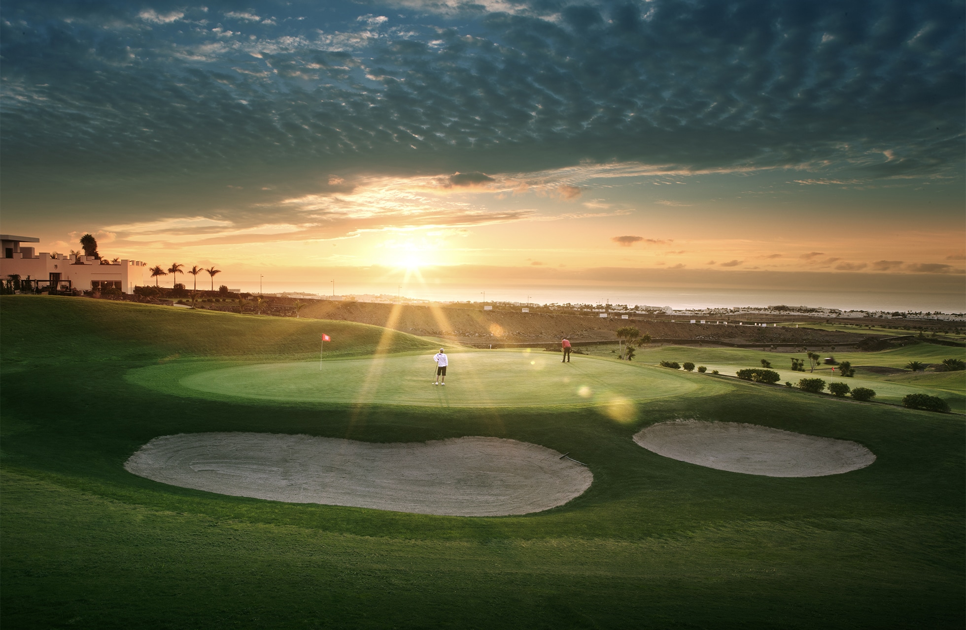 vidsträckt golfgreen med två bunkrar i förgrunden och flagga på hålet. flera golfspelare syns längre bort på banan. solen står lågt över horisonten och lyser över öppet landskap med havet i bakgrunden.
