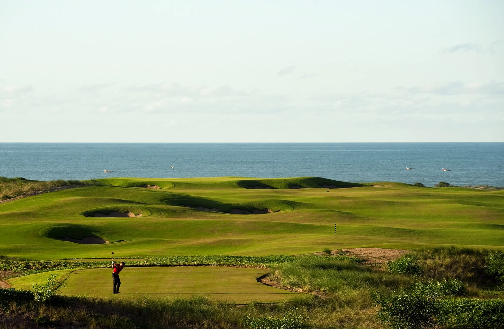 vidstrakt golfbane i kupert terreng med grønne fairwayer og greener, plassert ved havet. en golfspiller er synlig på banen med havet i bakgrunnen.