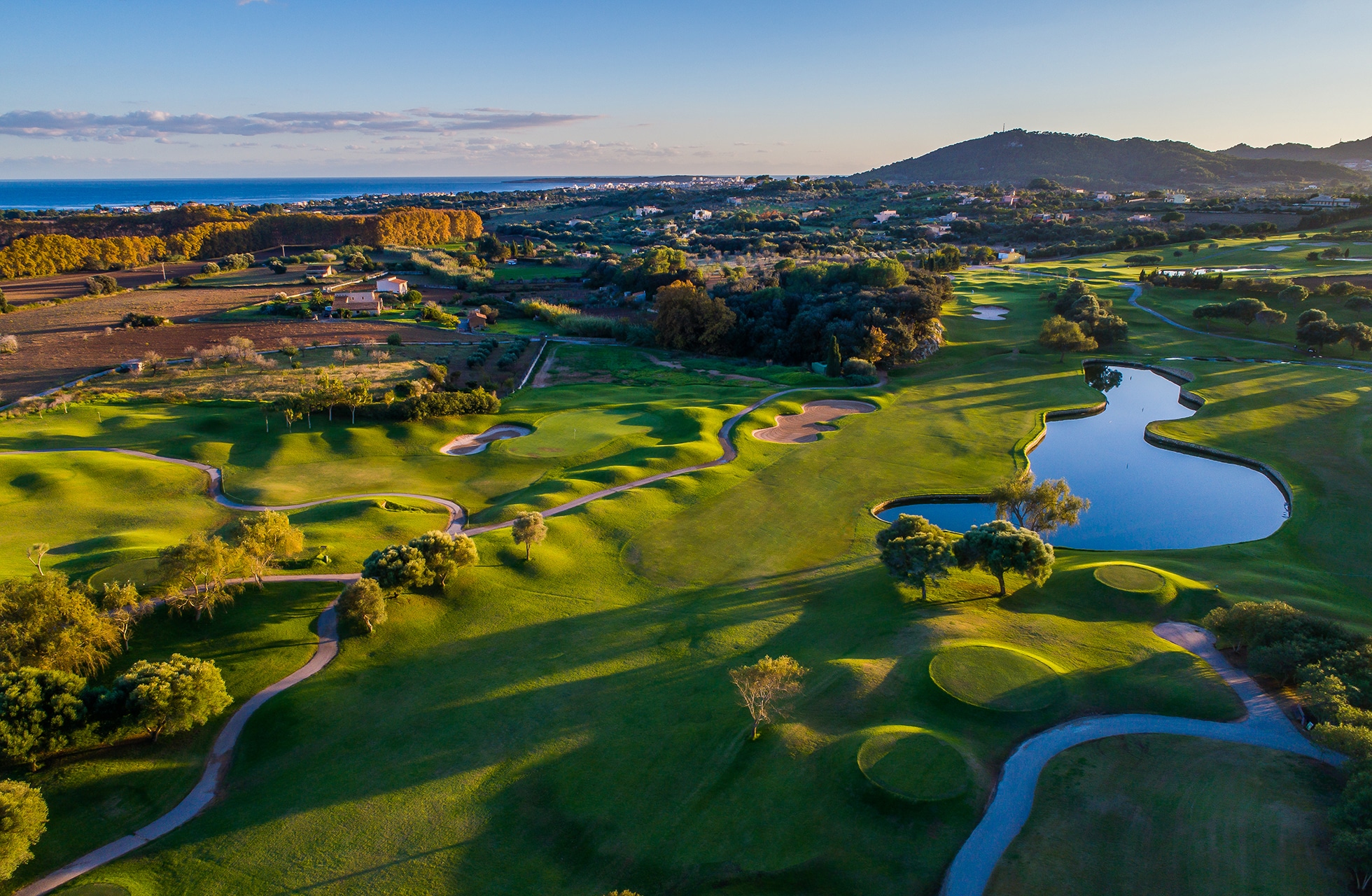 ilmakuva pula golf resortista mallorcalla, jossa näkyy kumpuilevia väyliä, viheriöitä ja suuri vesialue avoimessa maisemassa lähellä rannikkoa.