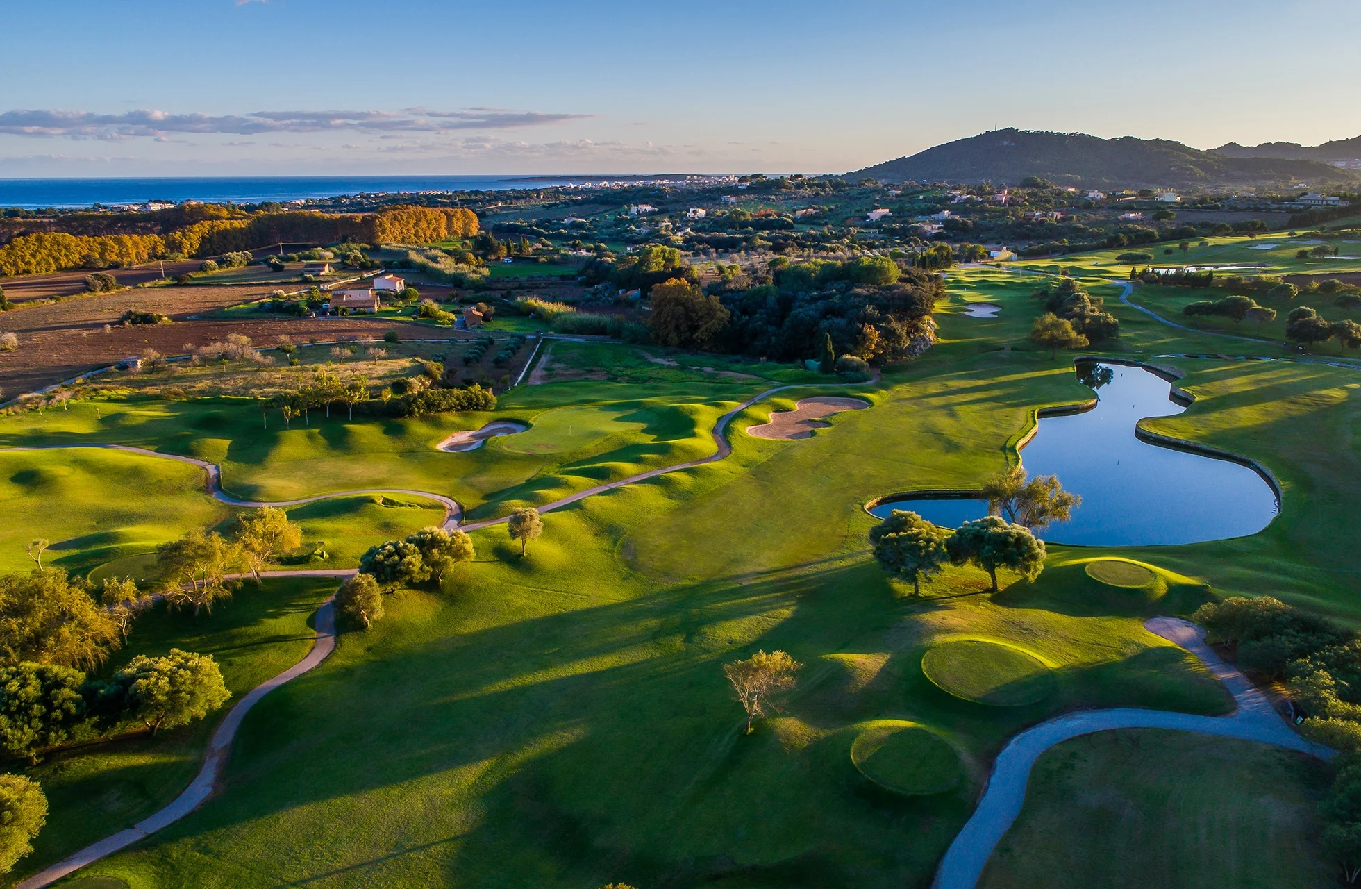 ilmakuva pula golf resortista mallorcalla, jossa näkyy kumpuilevia väyliä, viheriöitä ja suuri vesialue avoimessa maisemassa lähellä rannikkoa.