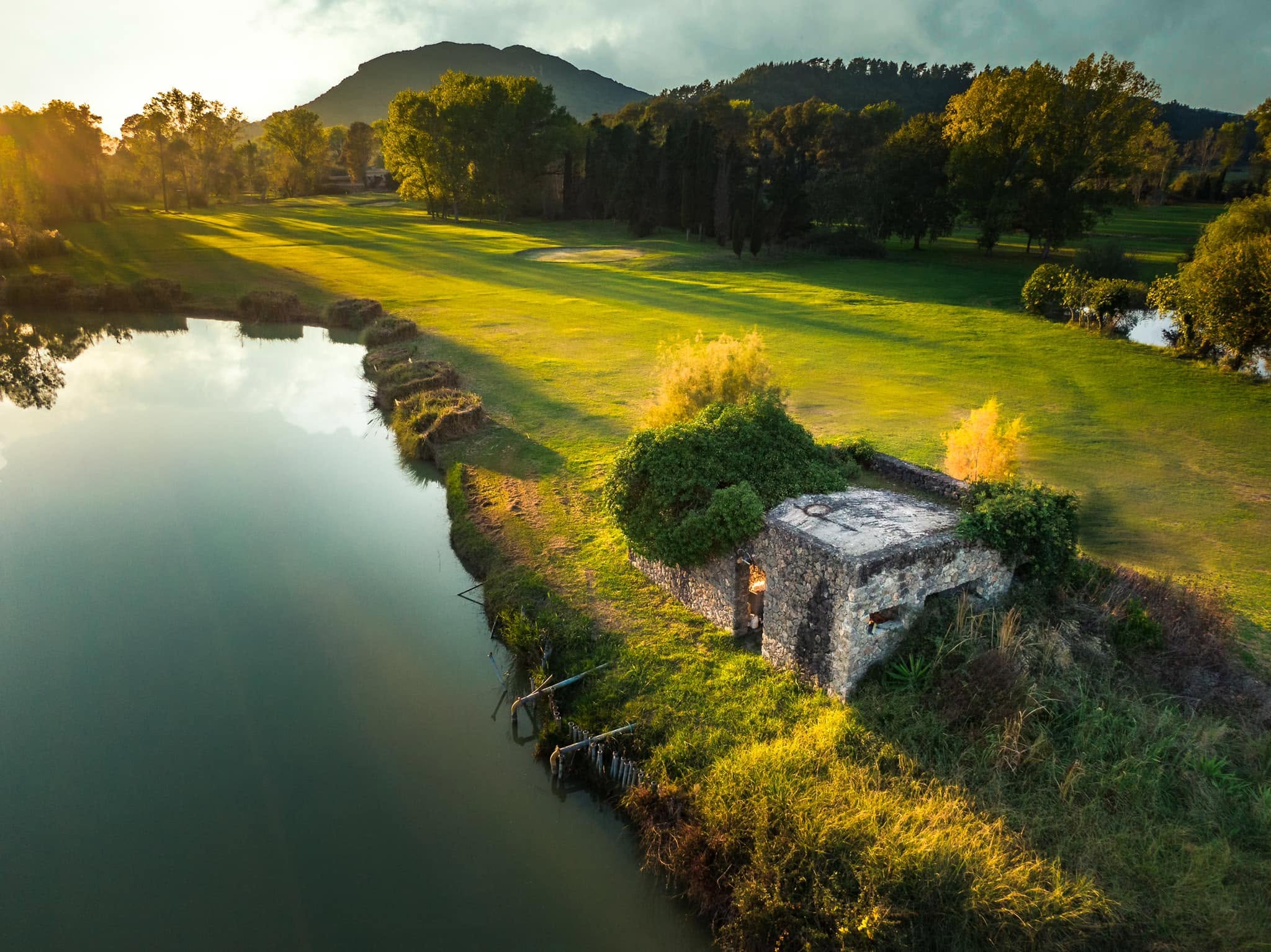 golfbana vid vattenkant med liten stenbyggnad och fairway i varmt kvällsljus