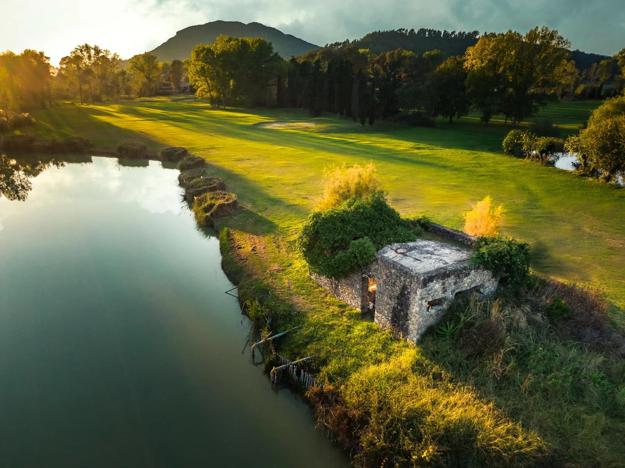 golfbana vid vattenkant med liten stenbyggnad och fairway i varmt kvällsljus