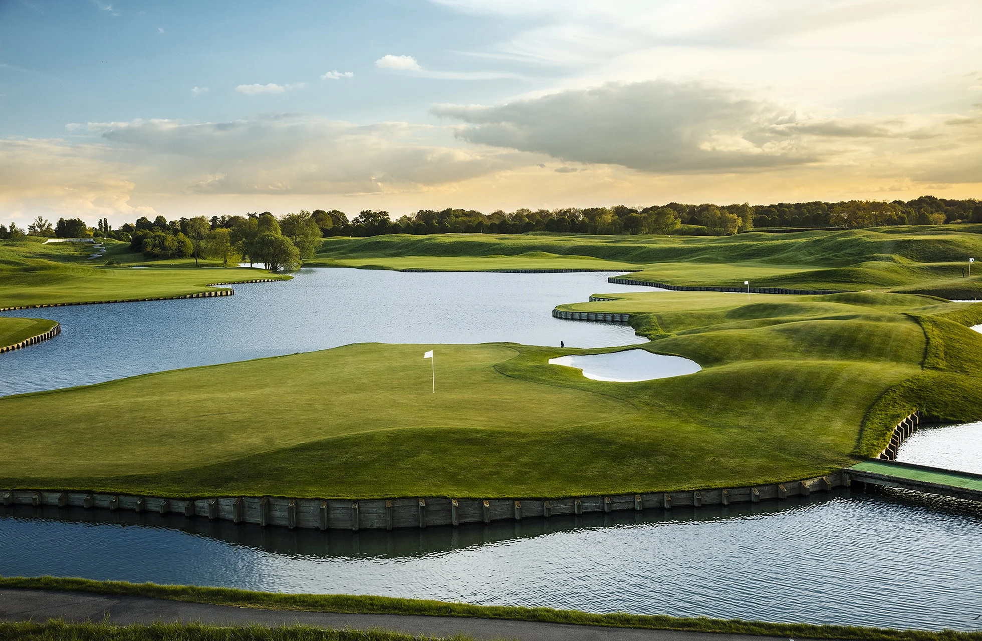 velstelt golfgreen omgitt av vann og bunkere i bølgende landskap, med flagg på greenen og åpne fairwayer i bakgrunnen