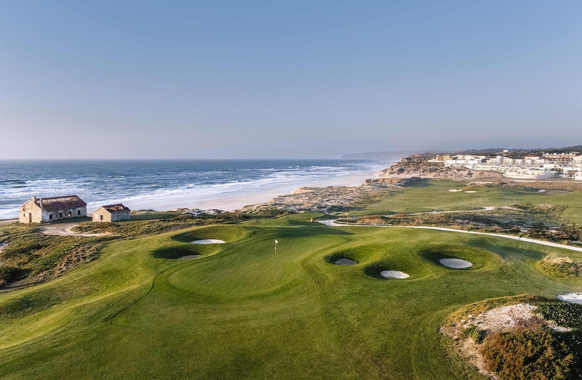 Lissabon_Praia-degolfgreen med flere bunkere i forgrunnen, omgitt av kortklipte fairwayer og naturlig vegetasjon, med havet og kystlinjen synlig i bakgrunnen i klart dagslys-rey_golf