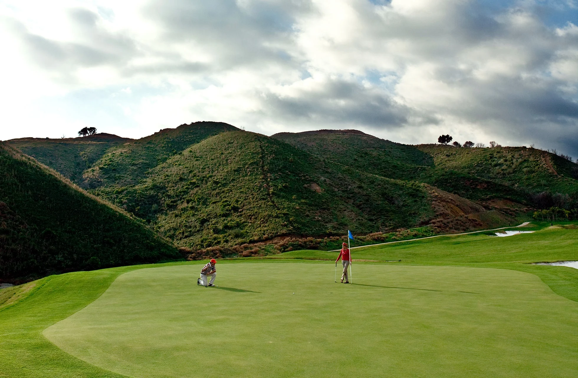 golfgreen på villa padierna golf med flagg og to golfspillere som putter, omgitt av grønne åser og fjell i mykt dagslys