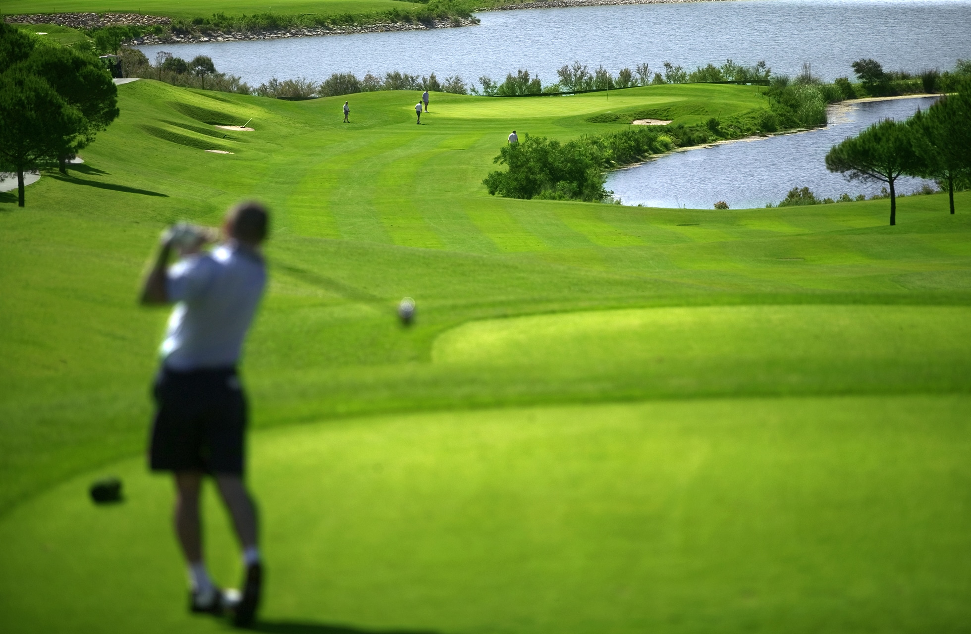 golfspiller i sving på tee med fairway og vannhindre i grønt landskap, greener og flagg i bakgrunnen